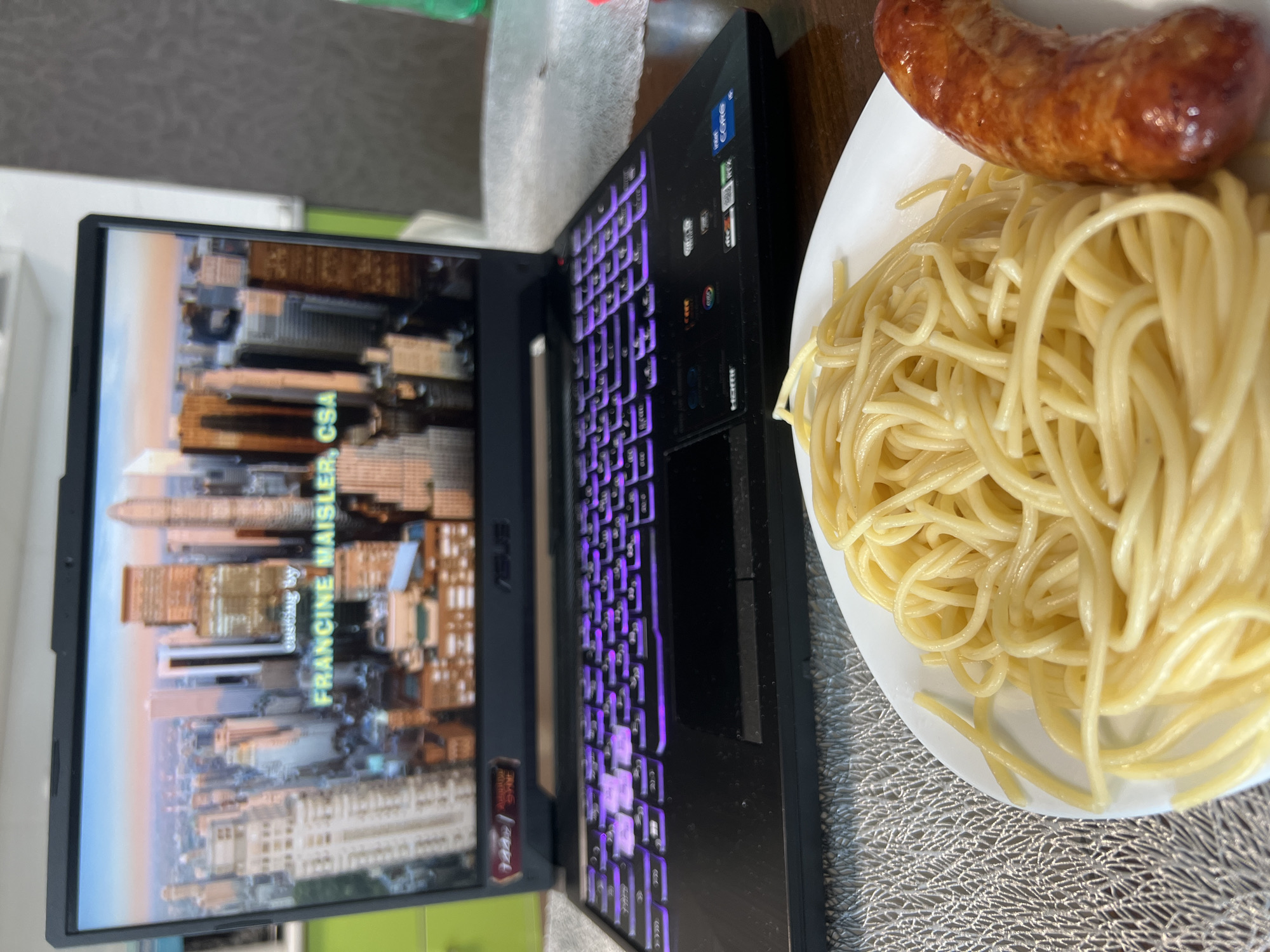 A black ASUS laptop with a brightly lit purple keyboard is to the left of a white plate of spaghetti and a sausage. The laptop screen displays a cityscape with the words "FRANCINE MAISLER, CSA." The setting appears to be indoors on a gray textured surface. The photograph does not show the city of Krasnoyarsk, Russia.