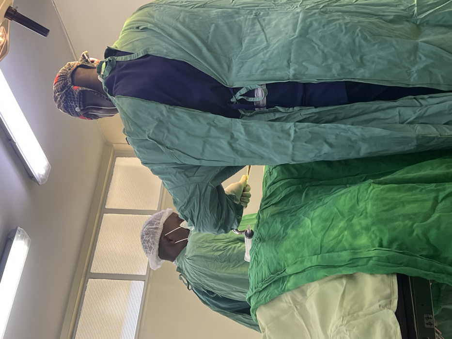 The scene depicts an operating room within a medical facility in Nairobi, Kenya. Two African males, identifiable as medical professionals, are engaged in a surgical procedure over a patient.
The individual in the foreground, positioned with his back to the viewer, wears a green surgical gown, open at the back, revealing a dark blue scrub top and trousers. A plaid surgical cap covers his head. His right hand, gloved in a light beige material, holds a gold-colored medical instrument positioned above the operating table.
To the left, another medical professional is shown in profile, looking down at the surgical field. This individual wears a green scrub top, a white disposable surgical cap, a white surgical mask covering the mouth and nose, and light beige gloves. His gloved hands hold a transparent syringe or suction device with a white plunger and grey tip, also positioned over the patient.
The patient lies supine on a medical operating table, completely covered by layered green surgical drapes. The top layer is a darker, crumpled green, while a lighter green drape is visible beneath it.
The environment features white walls and a white ceiling. Two rectangular fluorescent light fixtures are mounted on the ceiling, providing bright illumination. A portion of a specialized surgical lamp arm is visible on the upper right. Upper sections of the walls contain several horizontal rectangular windows fitted with horizontal blinds, suggesting an exterior wall for privacy or light control.