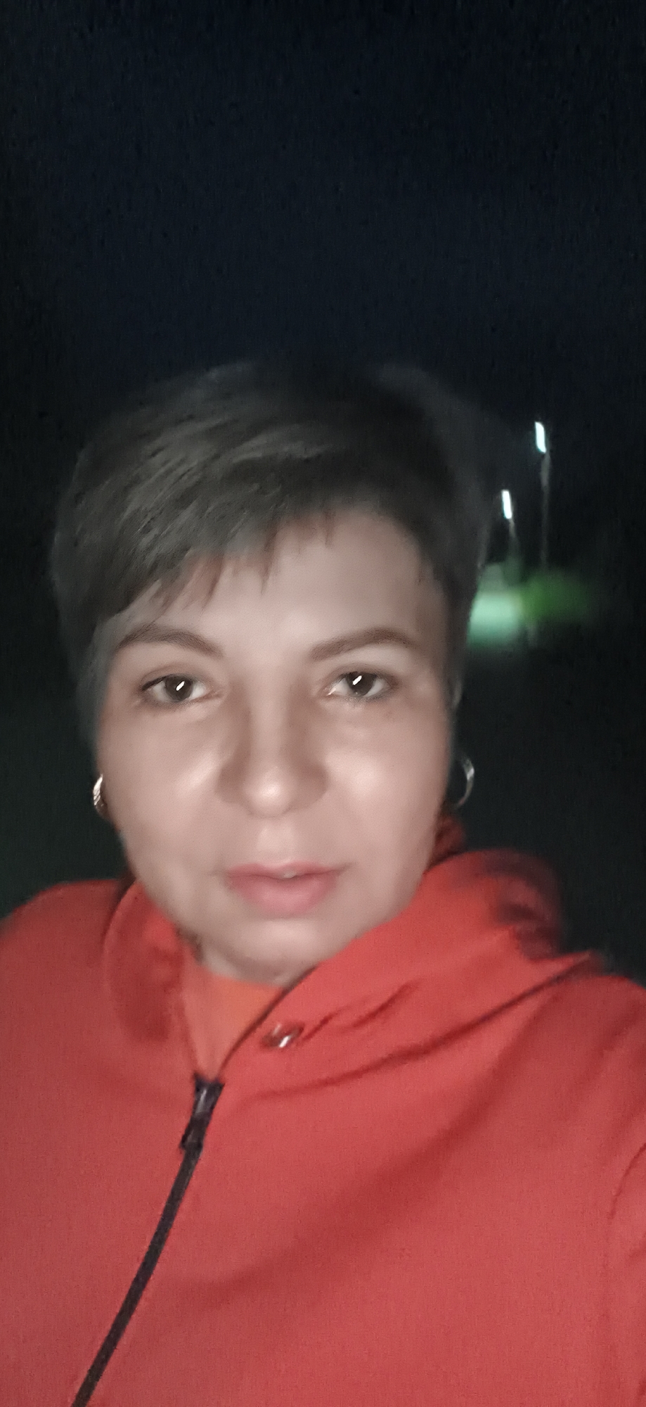 A medium close-up shot shows a woman with short, dark hair and light skin, facing the camera. She is wearing an orange hooded jacket with a zipper and an orange undershirt. She has on hoop earrings. Her lips are slightly parted. The background is dark, with some blurry lights and greenery visible, suggesting an outdoor setting at night. The image appears to be a selfie taken in the city of Fokino, Russia.