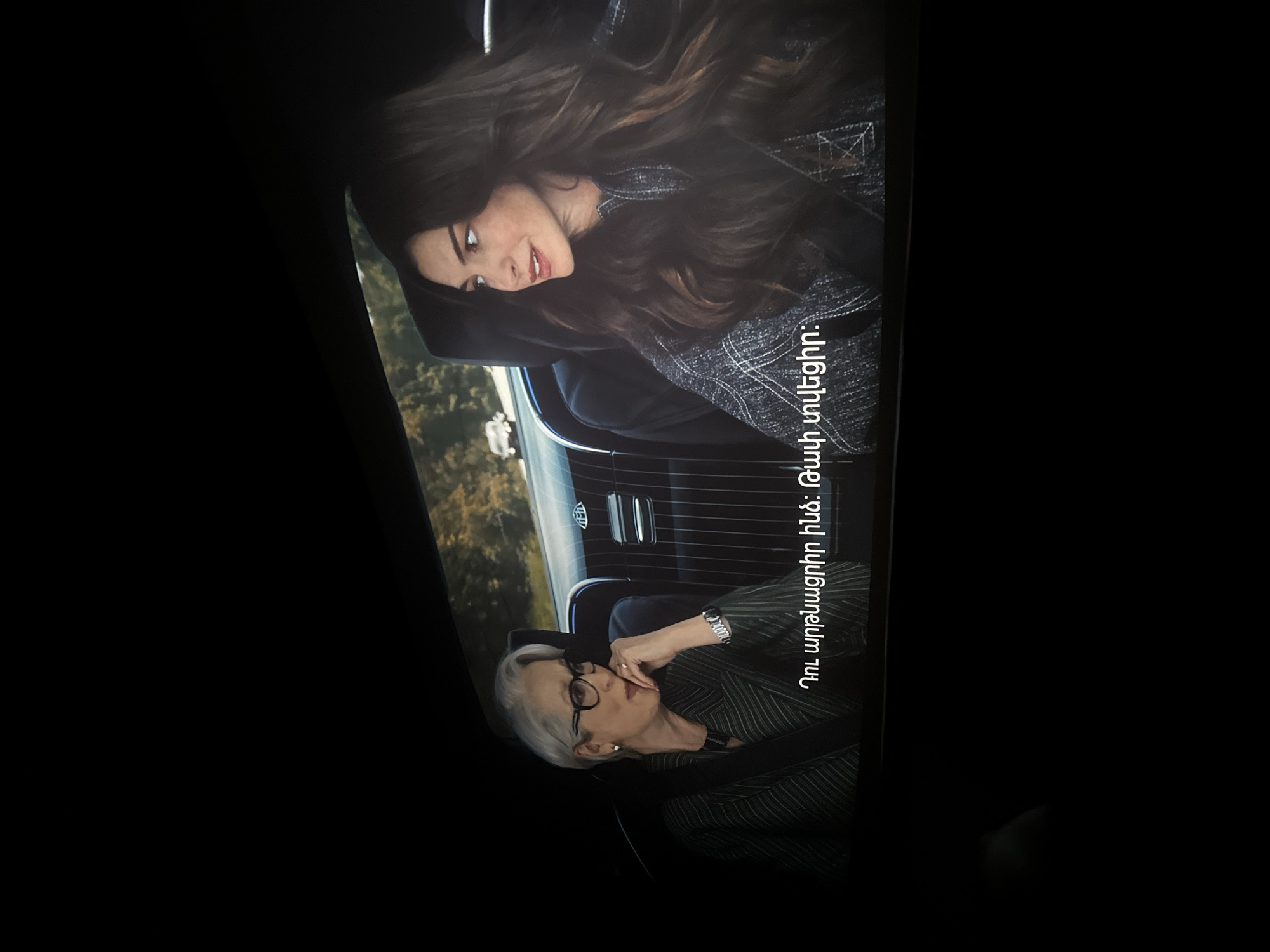 The image shows two women in a car. The woman on the left has short white hair, glasses, and is wearing a green and black striped blazer. She has her hand on her face. The woman on the right has long brown hair and is wearing a grey jacket. They appear to be in the back seat of a car, with the outside view showing a grassy area with a building. Text in an unknown language is visible in the center. There is no information indicating the location is in Yerevan, Armenia.