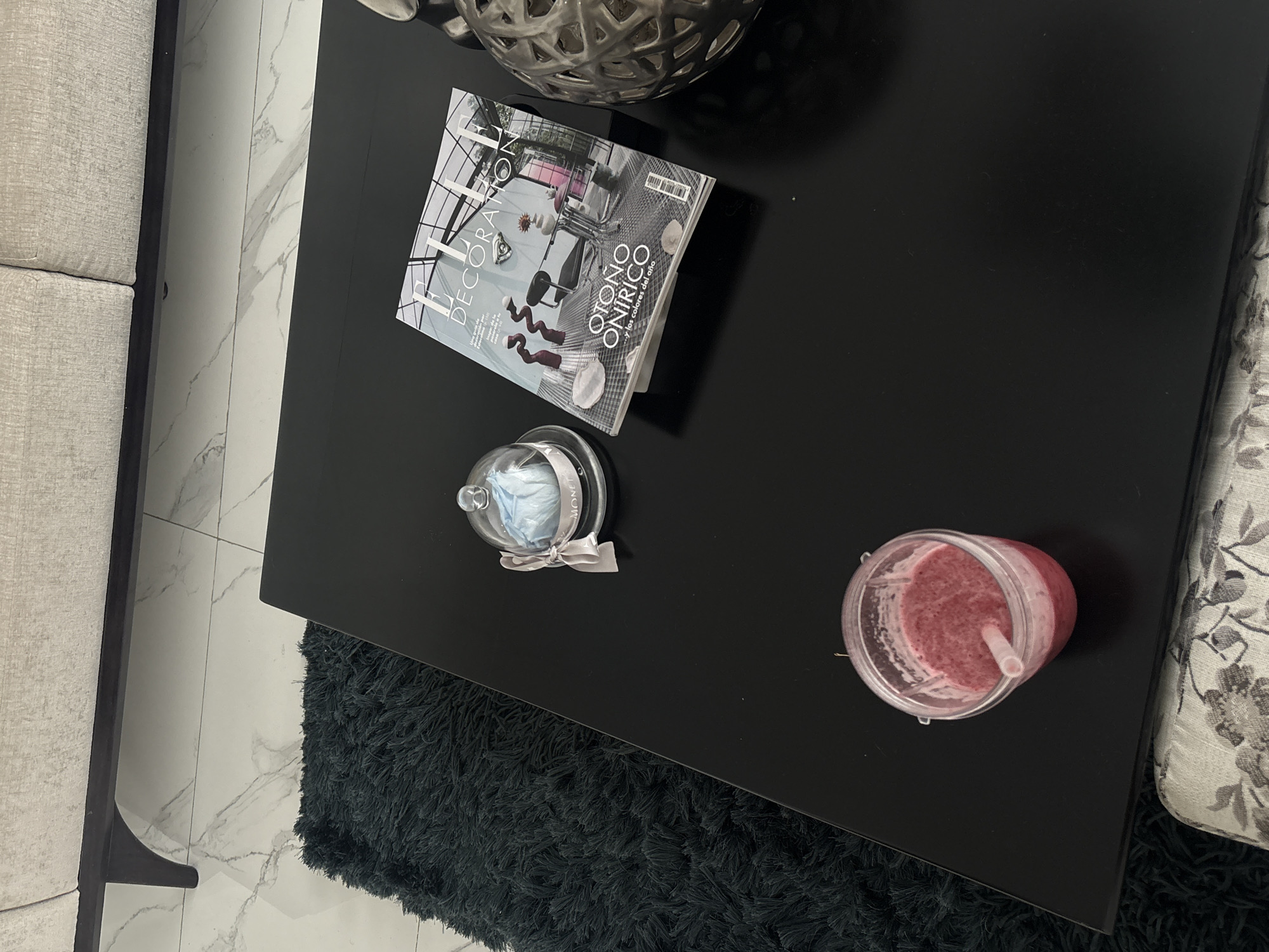 A high-angle shot depicts a black coffee table with several items. The table is situated partially on a dark gray, shaggy rug. On the table, from top-left to bottom-right, there is an ornamental silver spherical object, a magazine titled "Elle Decoracion" (Spanish Edition) with the words "Otono Onirico" visible, a small glass cloche with a white ribbon tied around it, and a clear plastic cup containing a pink smoothie with a straw. The edge of a light gray sofa is visible on the left, and the corner of a patterned sofa is visible on the right. The floor is tiled in a marble pattern.
This image does not contain information to determine the location in San Bernardino Tlaxcalancingo, Mexico.