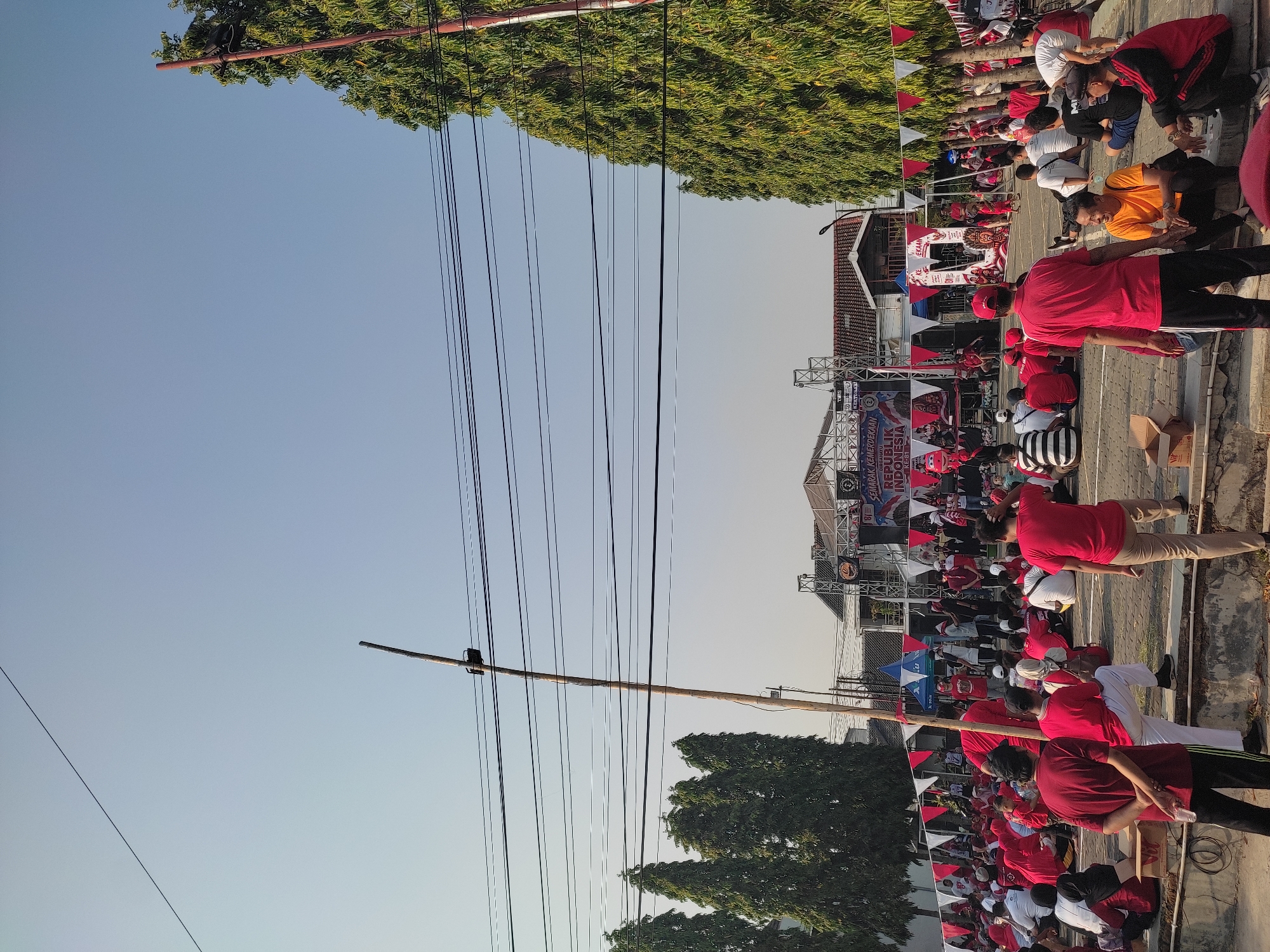 The image depicts an outdoor gathering in what appears to be a public space in the city of Ponorogo, Indonesia. A large crowd, predominantly dressed in red and white, is seated on the ground facing a stage. The stage is set up with a large banner that reads "SEMARAK KEMERDEKAAN REPUBLIK INDONESIA" and "81 DIRGAHAYU REPUBLIK INDONESIA". Several people are standing on the stage, and some appear to be addressing the crowd. Decorative triangular flags in red and white are strung across the area. Tall green trees line the background, and utility poles with multiple wires are visible overhead. The ground is paved with cobblestones. Several individuals in the foreground are also seated or standing, some interacting with each other. The overall atmosphere suggests a celebratory event, possibly related to Indonesian Independence Day.