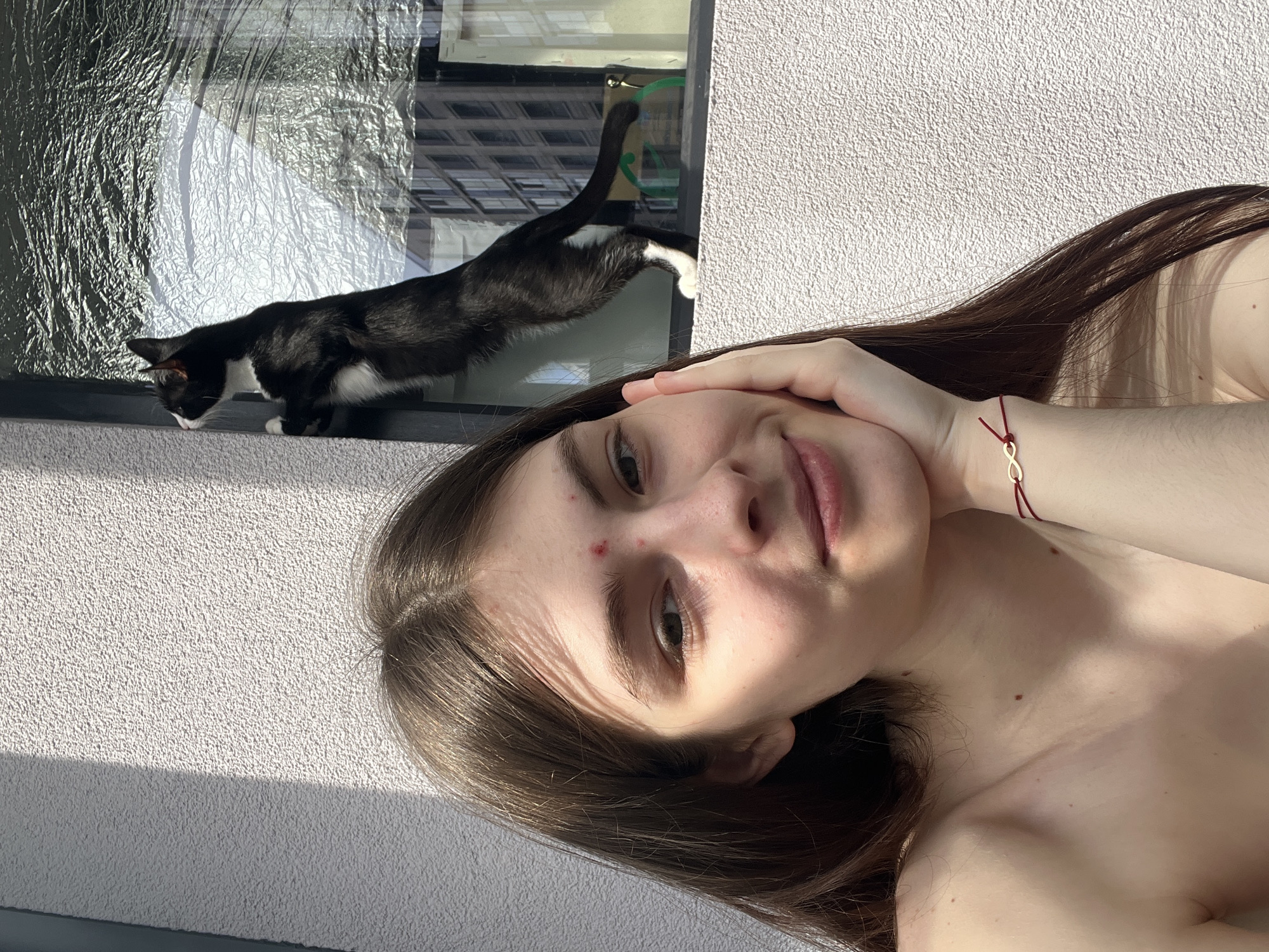 The image, taken in Saint Petersburg, Russia, captures a selfie-style portrait of a young woman alongside a black and white cat. The scene is set outdoors, featuring a textured white stucco wall and a window.
In the foreground, a young woman with long, dark brown hair is positioned diagonally, looking towards the camera with a neutral to slightly smiling expression. Her left hand is raised, partially covering her cheek and supporting her head, while her right arm, adorned with a red string bracelet featuring a gold-colored infinity symbol, is extended out of frame, likely holding the camera. Her upper torso appears bare, and her skin is illuminated by direct sunlight, which also casts subtle shadows. A couple of small reddish blemishes are visible on her forehead.
Above and behind the woman, a black and white tuxedo cat is seen on a narrow ledge directly below a window. The cat's head is lowered, appearing to sniff or look at something on the ledge, while its hindquarters and tail are partially visible, with its tail reflected in the window pane. The window is covered with a highly reflective, crinkled material, possibly foil or a textured film, which reflects surrounding urban buildings. A small green object, resembling a hanger or hook, is faintly visible inside the window.
The time of day is clearly daylight, indicated by the strong, direct sunlight casting crisp shadows on the textured white wall. No visible text is present in the image.