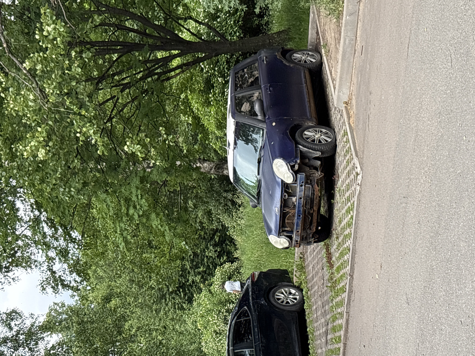 The image captures a blue hatchback car, appearing damaged and possibly abandoned, parked in a designated spot in Saint Petersburg, Russia. The car's front bumper and grill are missing, revealing rusty metal components. It has white accents on the roof. Beside it, a black sedan is parked. In the background, a person in a white shirt is walking away from the camera, heading towards a lush green area with trees and bushes. The ground is a mix of paved surface and what appears to be a grid-like eco-paver system. The overall environment suggests an urban or suburban setting with abundant greenery.
