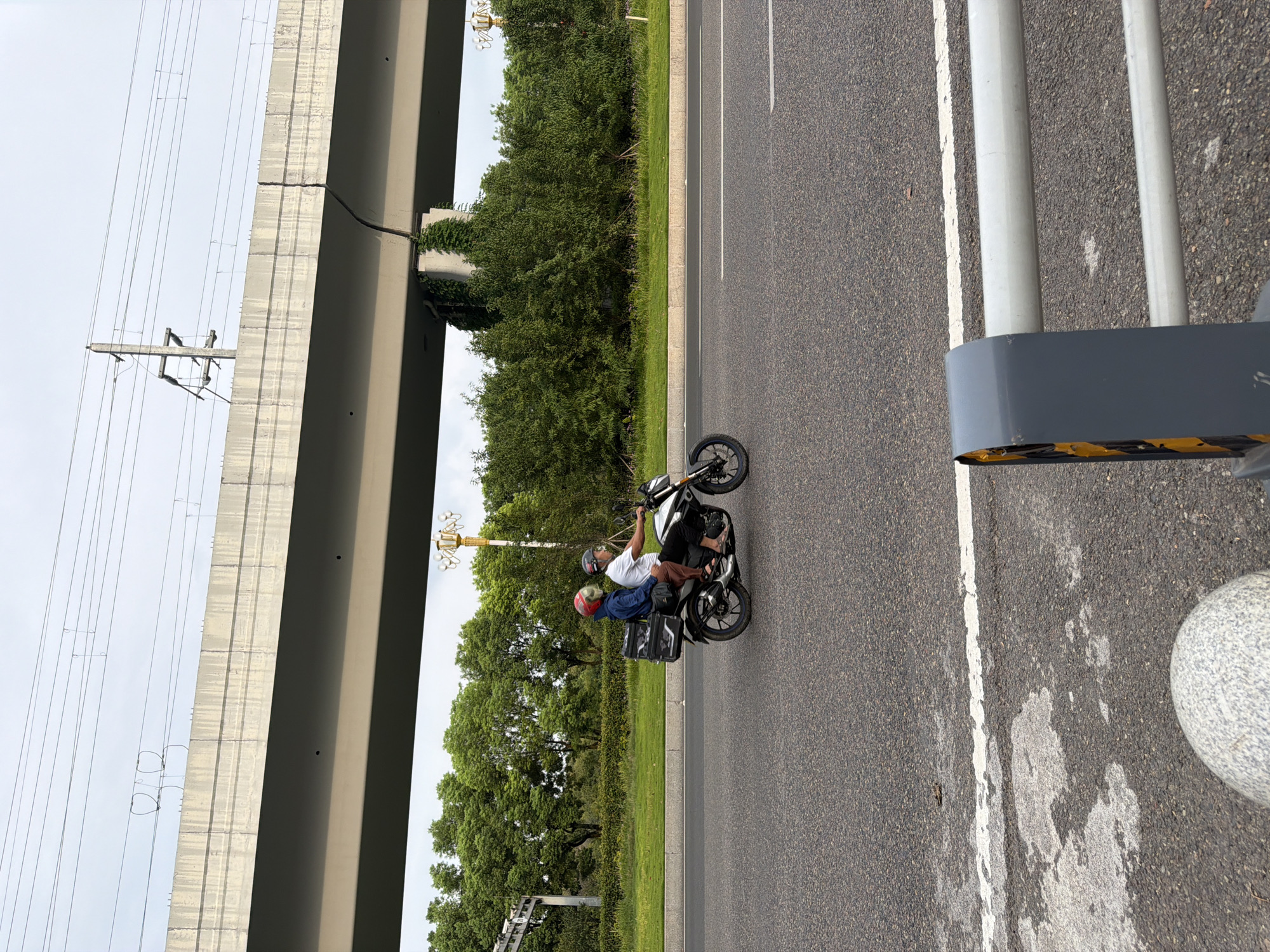 A motorcycle with two occupants, a driver and a passenger, travels on a paved road in Jiangjin, China. The driver, wearing a white t-shirt and black pants, operates the motorcycle. The passenger, in a blue jacket and brown pants, sits behind the driver. Both individuals wear helmets, one red and one gray. A black storage box is attached to the rear of the motorcycle. The road is flanked by a grassy area and lush green trees. Above the trees, a large elevated concrete structure, possibly an overpass or bridge, spans the scene. Overhead, power lines are visible against a cloudy sky. In the foreground, a portion of a grey barrier with yellow markings is visible.