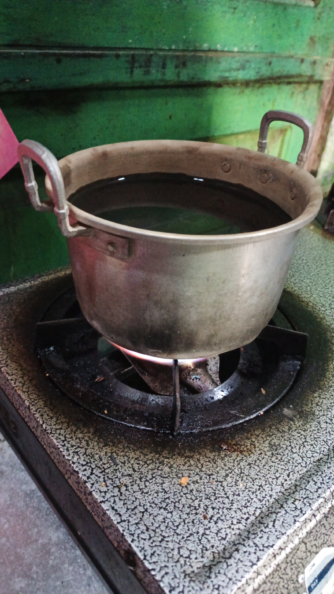 A metallic pot filled with water is placed on a gas stove in a kitchen. The pot has two handles and is slightly tarnished. The stove has a speckled gray surface and a black grate. A flame is visible beneath the pot, indicating it is being heated. The background is a green, textured surface, possibly a wall or cabinet. The scene appears to be in the city of Gunungreja, Indonesia.