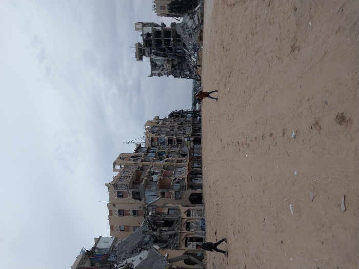 Two children walk on a sandy terrain next to damaged buildings. One child is near the top of the frame, and another is near the bottom. Debris and rubble surround the buildings, which are heavily damaged with exposed interiors. The sky is overcast.
