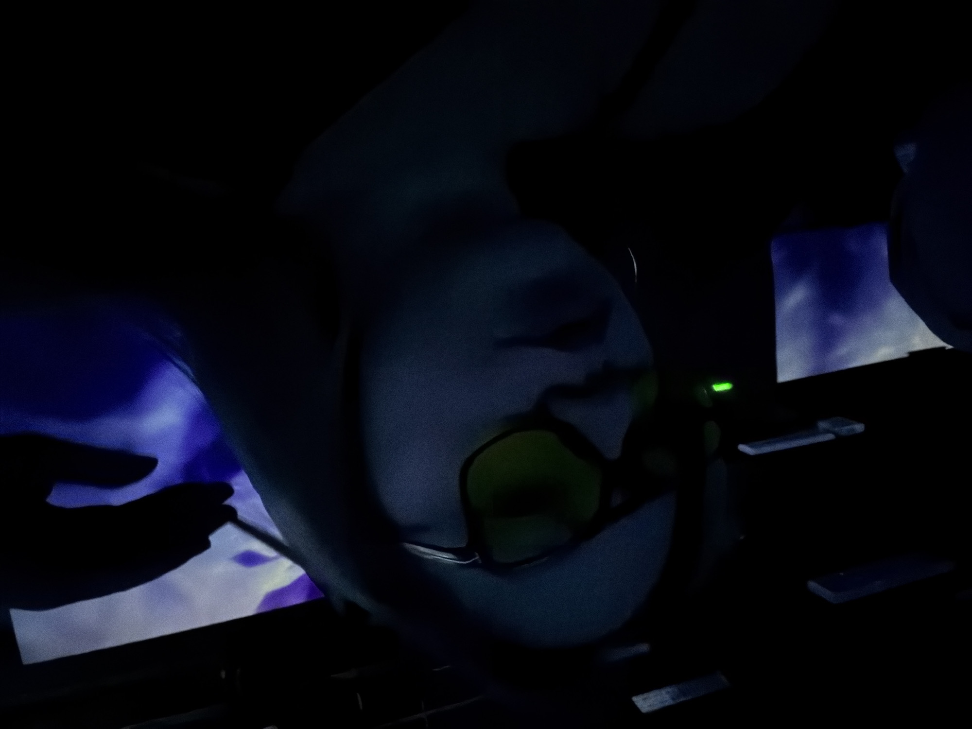 The image shows a close-up, upside-down view of a person wearing glasses with yellow lenses. The person's face is angled downwards, with their forehead and the top of their head visible, along with the bridge of their nose and eyes behind the glasses. The scene is dimly lit, creating a dark and somewhat mysterious atmosphere. To the left of the person, a shadowy hand is visible, reaching out towards a screen or surface displaying a blue and purple abstract pattern, possibly clouds or a digital display. To the right, the dark background features several flat, rectangular objects, some of which appear to be electronic components or screens. A small green light is also visible in the upper right portion of the image. The overall impression is one of being in a technologically advanced or futuristic environment, possibly a studio or a control room, with the low light and unusual perspective contributing to an enigmatic mood. No text is visible in the image. The time of day is indeterminate due to the artificial lighting, and there are no clear weather or specific location cues beyond the provided context of Moscow, Russia.