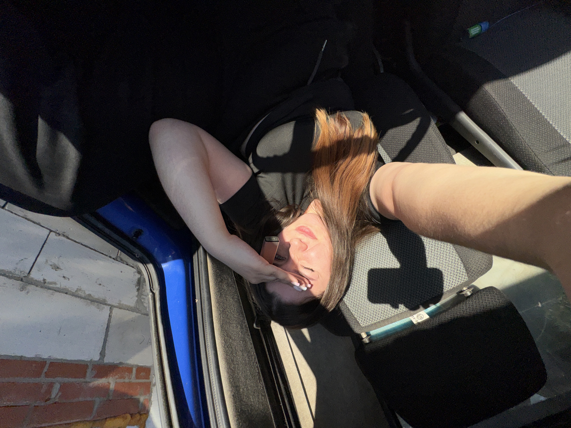 In Krasnokamsk, Russia, a woman is seen taking a selfie inside what appears to be a vehicle. She is lying down, with her head resting on a dark seat. Sunlight casts a strong shadow across her face, and she is holding her hand to her forehead, possibly shielding her eyes from the bright light. She is wearing a black t-shirt and black pants. A pink vape pen is visible near her hand, and a small green and blue object is on the floor of the vehicle. The interior of the vehicle features dark grey fabric seats. Through the window, a brick wall and some lighter colored blocks are visible, suggesting an outdoor urban or industrial setting.