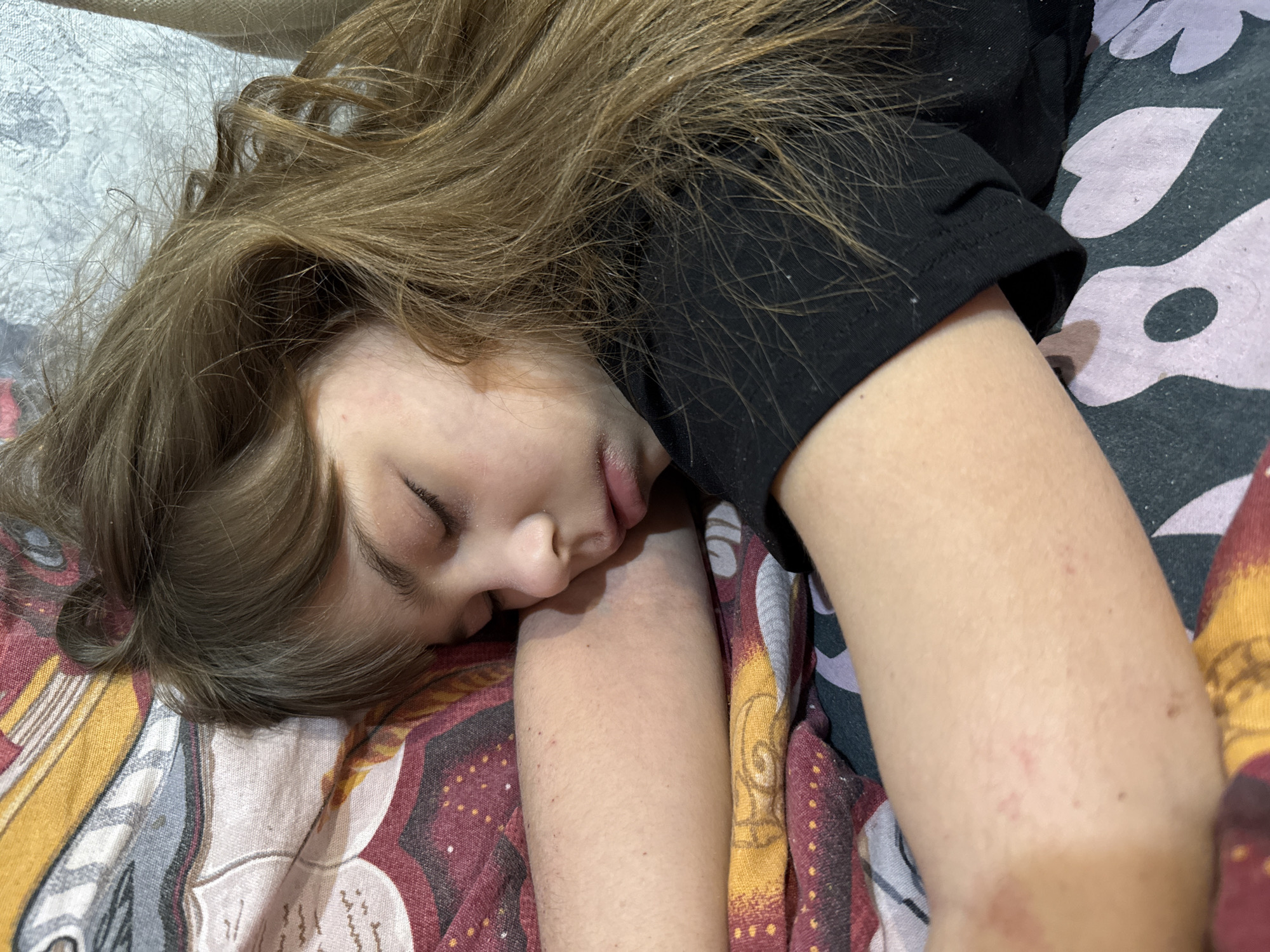 A young person with light brown hair is sleeping on a patterned bedspread, with a dark-colored t-shirt covering their arm. No identifiable location within Rusanovo, Russia, is discernible from the image. The scene depicts a person at rest.