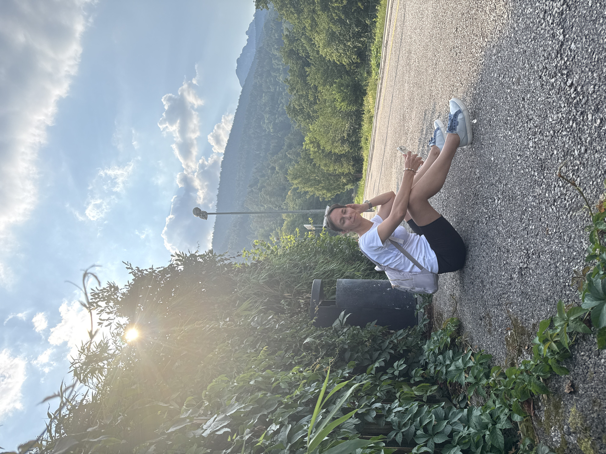 A woman is sitting on a paved road, leaning back with her arms behind her. She is wearing a white t-shirt, black shorts, and light blue sneakers. She has a white backpack on and is holding a phone in her hand. She is smiling and appears relaxed. The scene is a roadside with lush green trees and mountains in the background. The sun is shining, and the sky is blue with some clouds. The image was taken in Calceranica al Lago, Italy, likely in the late afternoon given the golden light. There is a trash can next to her. The road appears to be a rural road, possibly on a mountain pass given the elevation and foliage.
