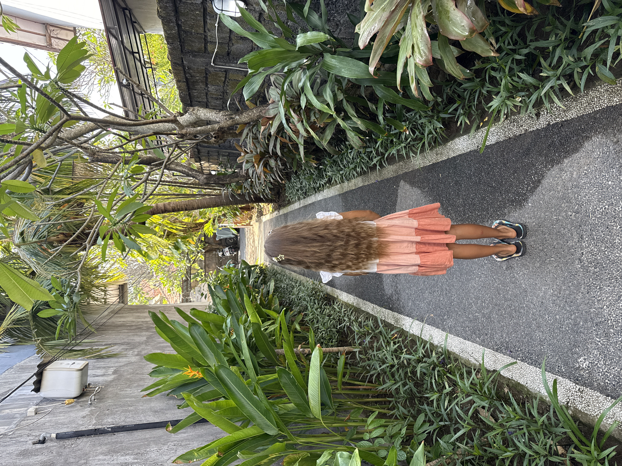 A young girl with long, wavy brown hair stands with her back to the camera, facing away down a narrow paved walkway. She is wearing a pink and orange tiered dress and black flip-flops. The walkway is flanked by lush green foliage and tropical plants. On the left, a textured grey wall runs parallel to the path, with a white electrical box mounted near the top. On the right, a darker stone wall is visible, with more plants growing alongside it. The scene suggests a tropical environment, possibly a courtyard or a residential alleyway in Denpasar, Indonesia, given the architectural style and vegetation. Sunlight filters through the trees, casting shadows on the walkway. In the distance, further down the path, what appears to be a motorcycle is parked.