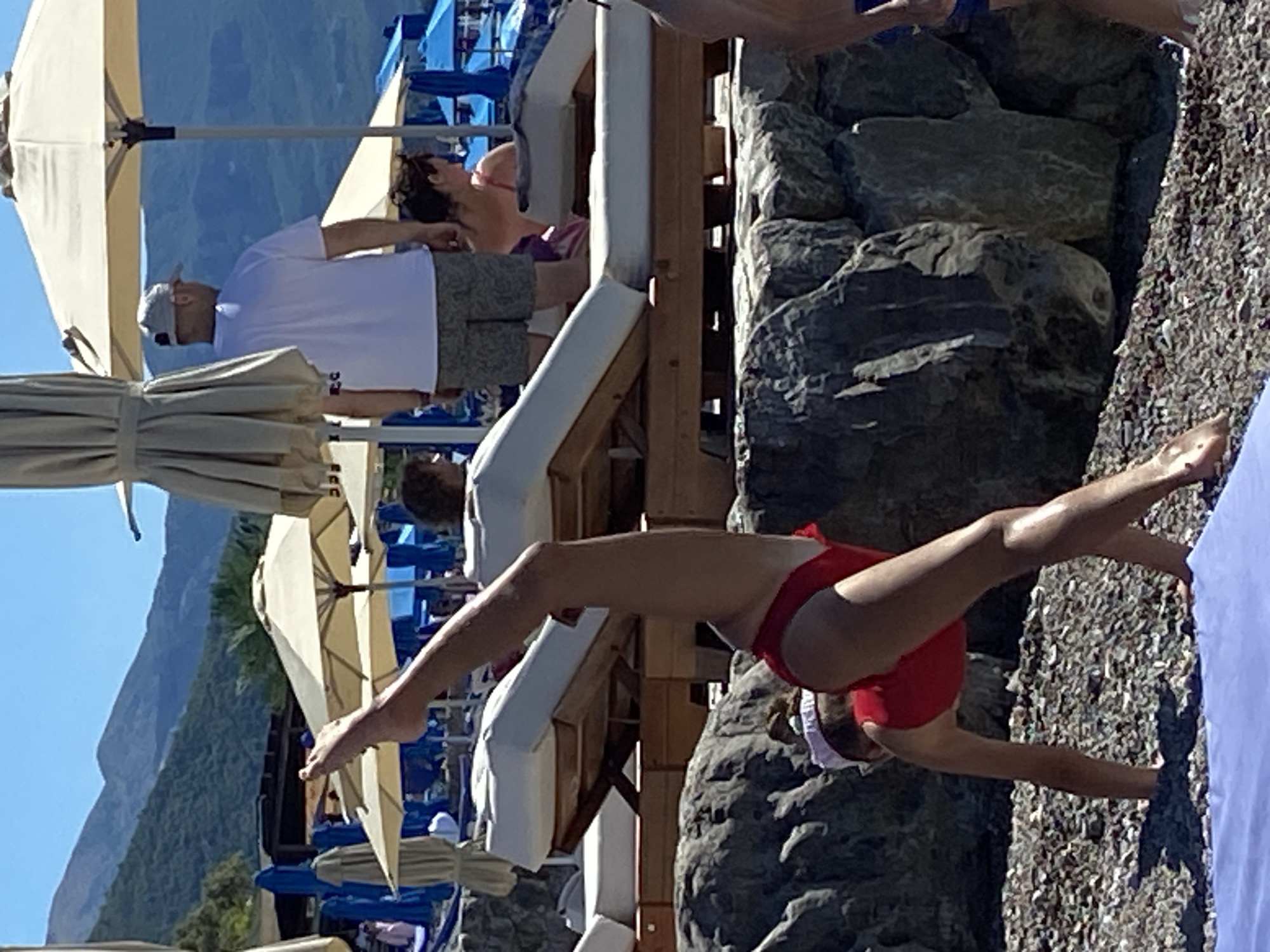 A young woman in a red swimsuit is performing a gymnastic pose on a pebble beach. Her body is arched, with her hands on the ground and her legs extended vertically upwards. She is wearing a white headband. In the background, there are several beach umbrellas, lounge chairs, and people. Mountains are visible in the distance under a clear blue sky. The scene appears to be at a beach resort in Prijevor, Montenegro.