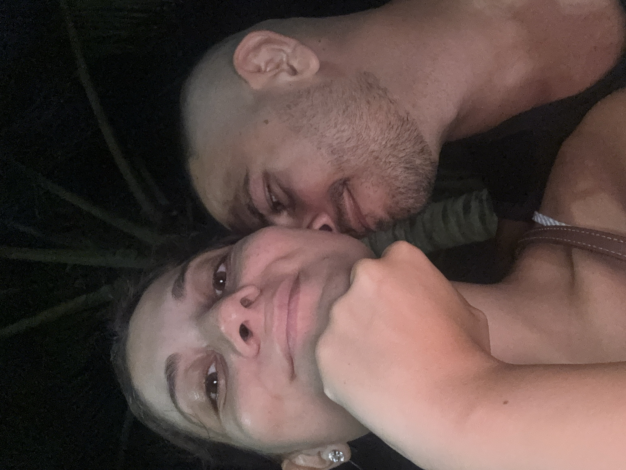 A close-up, vertically oriented photograph shows a woman and a man embracing. The woman, in the foreground, is fair-skinned and looks at the camera. Her right hand is near her face. She has dark hair pulled back, brown eyes, and a small earring is visible in her left ear. Behind her, a bald, bearded man embraces her. His eyes are closed. The background is dark. The location is unknown.