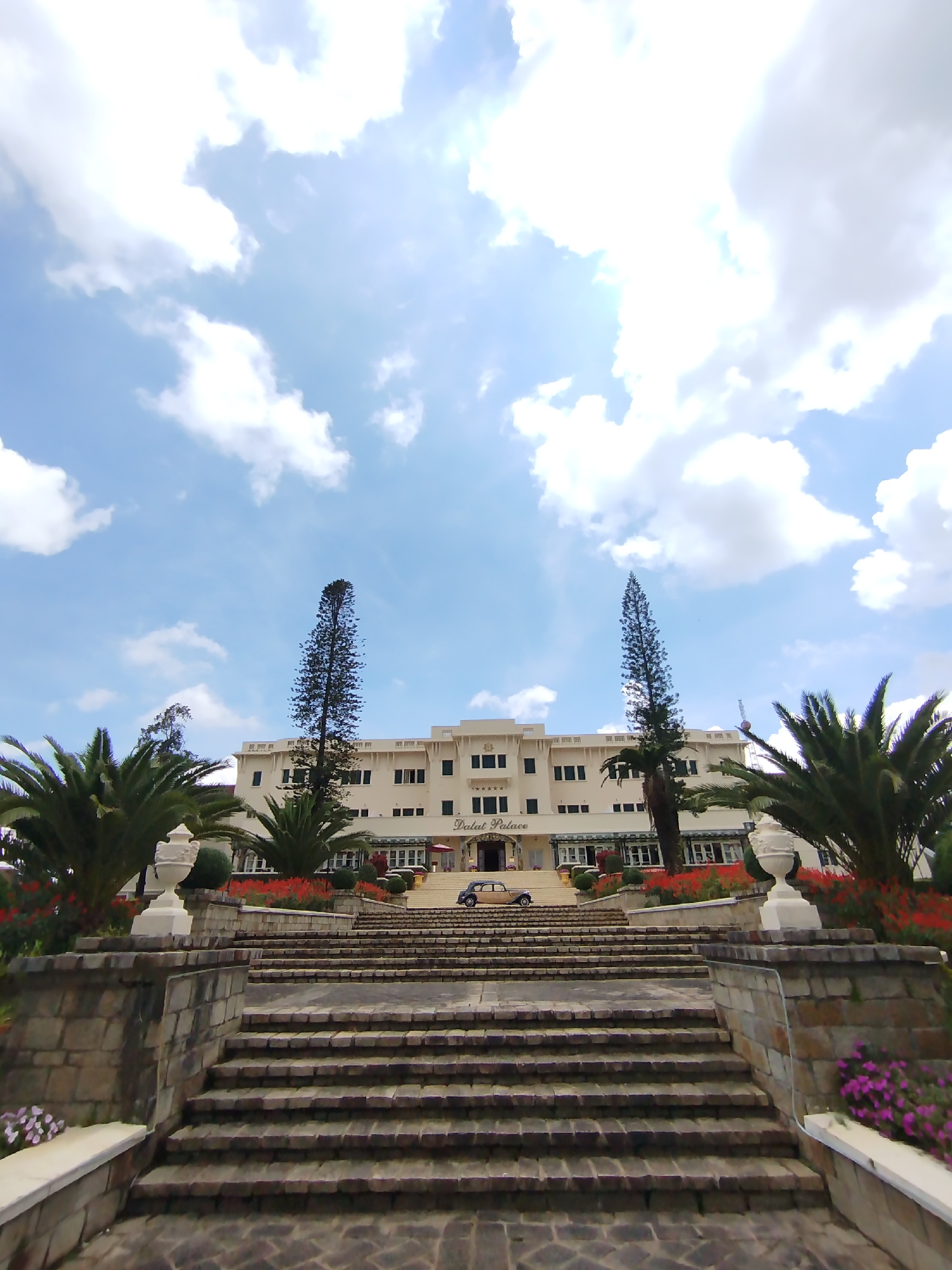 The image shows the Dalat Palace Hotel in Da Lat, Vietnam. The hotel is a large, light-colored building with multiple windows and balconies. It is situated at the top of a long, wide set of stone stairs. In front of the hotel, there are well-manicured gardens with red flowers, palm trees, and two tall, thin coniferous trees. A vintage car is parked in front of the hotel entrance. The sky is blue with scattered white clouds. The photograph is taken from a low angle, emphasizing the height of the staircase and the building. The overall scene suggests a historic and grand hotel setting.