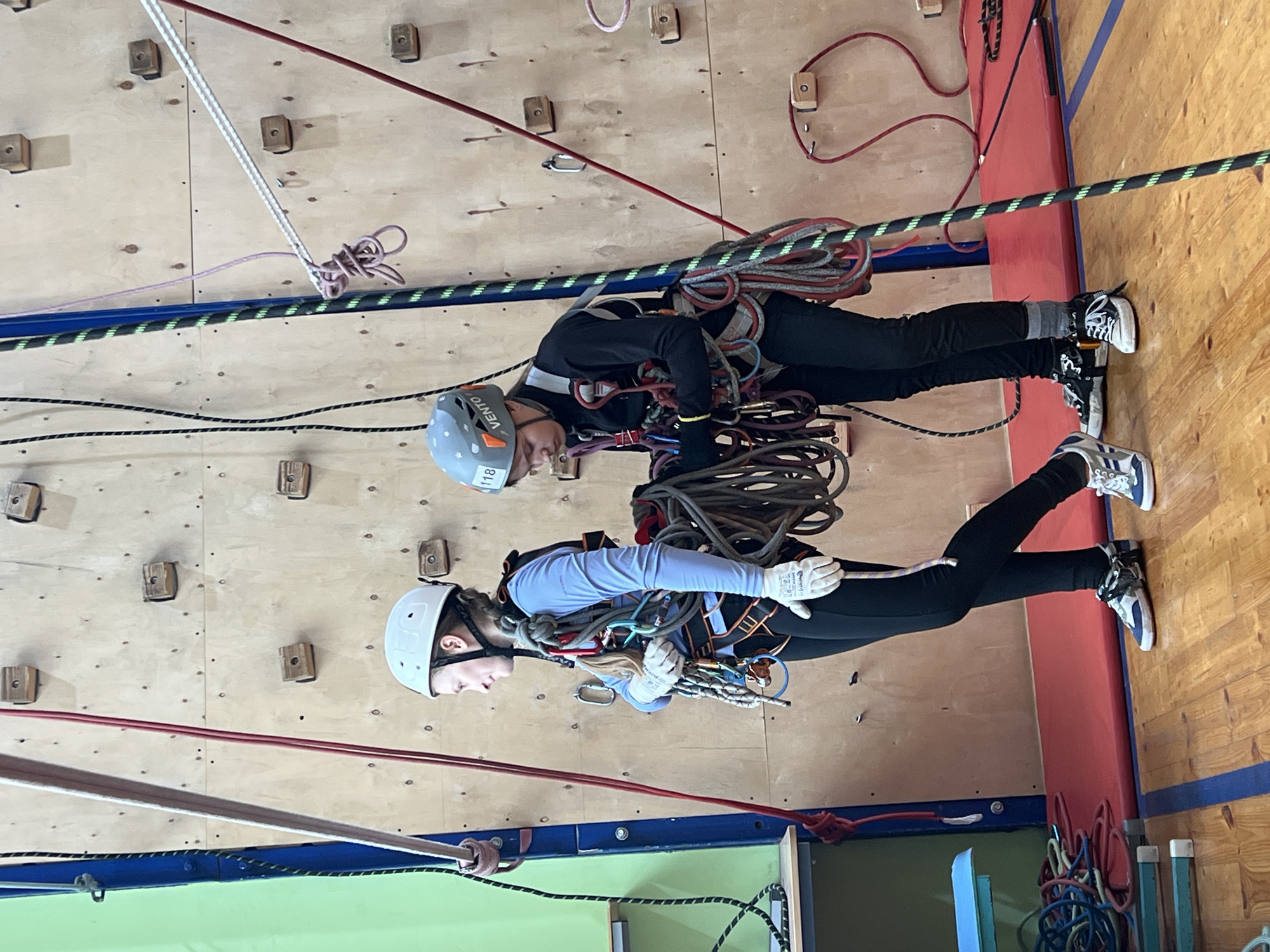 Two individuals are suspended by ropes and harnesses against an indoor climbing wall. The wall is made of wood and has various holds attached. Both individuals are wearing helmets and are equipped with climbing gear, including ropes and carabiners. The person in the foreground wears a light blue shirt and white gloves. The person in the background wears a black jacket. The climbing area has a blue and green base with red padding. The location is an indoor climbing facility, presumably in the city of Chaikovsky, Russia.