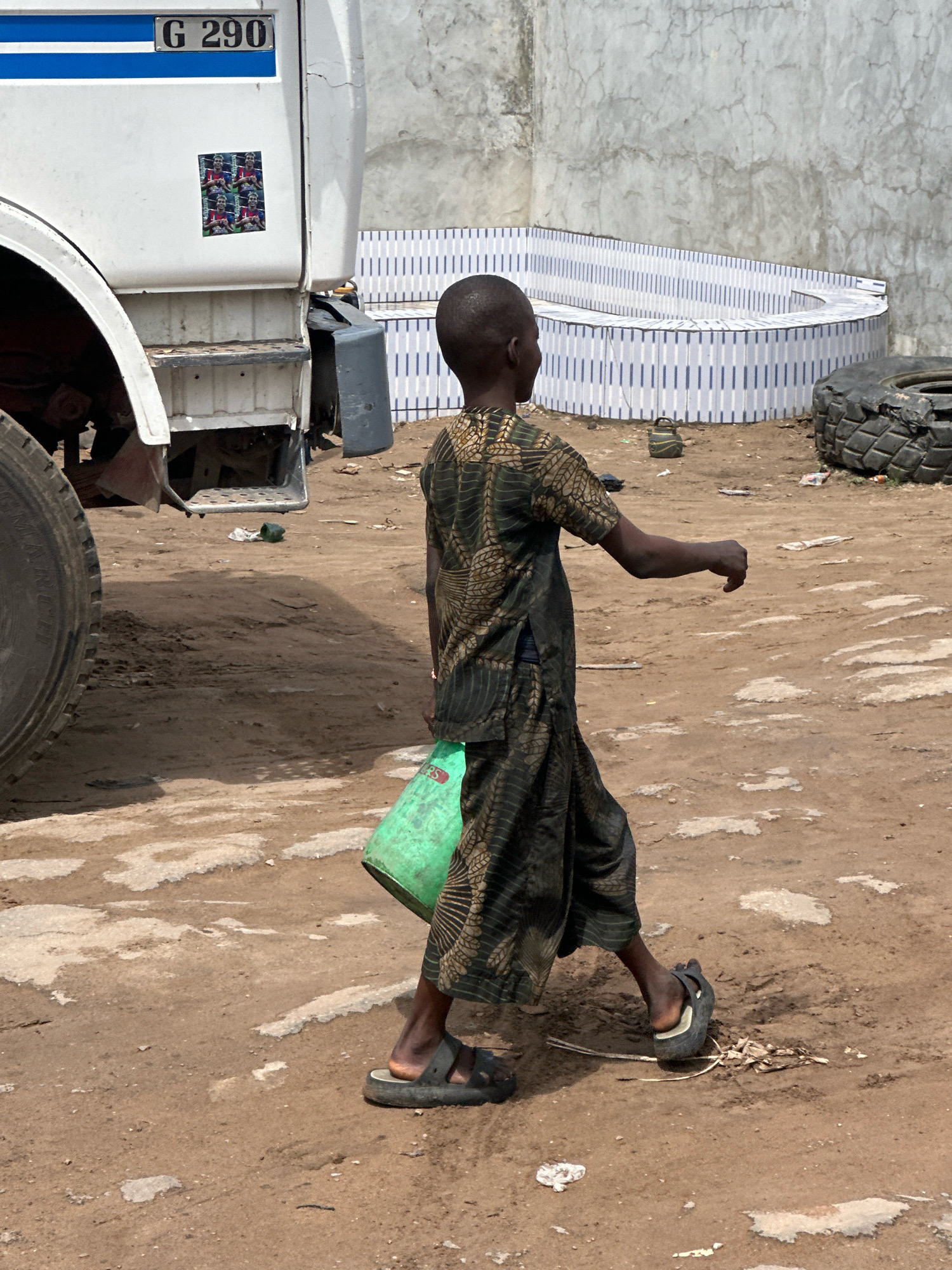 In Ajara, Nigeria, a young boy walks in profile to the right, carrying a green bucket. He wears a dark, patterned tunic and pants, and black sandals. He is walking on a dirt ground next to a white truck with blue stripes and the license plate "G 290." In the background, a tiled wall surrounds a circular structure, and a worn tire lies on the ground. The environment appears dry and dusty, with scattered debris.