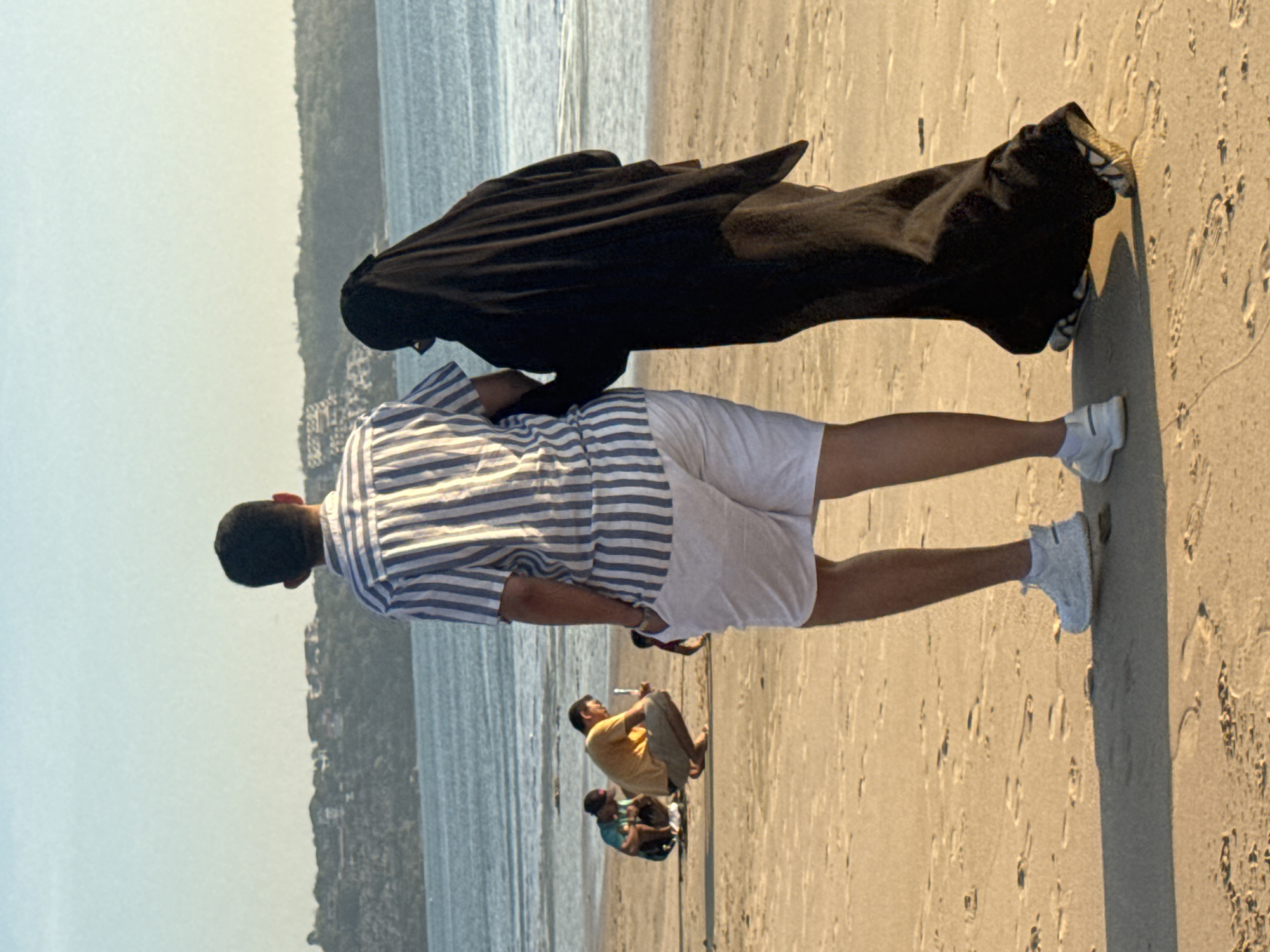The image shows a couple walking on a sandy beach in Kuta, Indonesia, with the ocean and a tree-covered hillside in the background. The man, on the left, is wearing a blue and white striped short-sleeved shirt and white shorts. The woman, on the right, is wearing a black abaya and hijab. They are walking away from the camera, towards the sea. In the mid-ground, two other individuals are crouched on the sand. One is wearing a yellow shirt and brown shorts, and the other is wearing a blue t-shirt and dark shorts, with a cap. The sand is marked with footprints. The lighting suggests it is late afternoon.