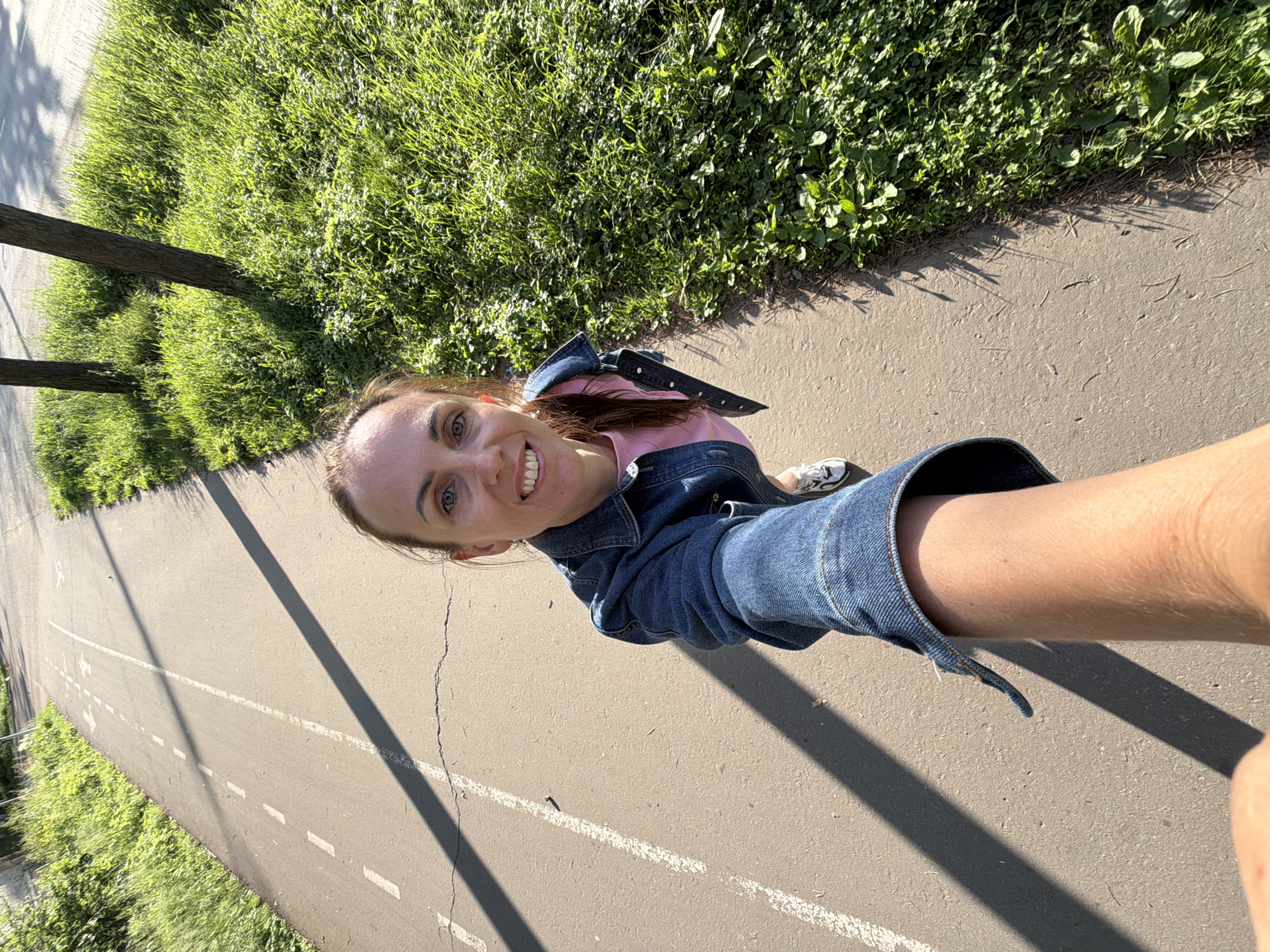 A woman takes a selfie with her arm extended forward. She is walking on a paved path with white lane markings, which appears to be a bike or pedestrian path. To her right is a grassy area with two trees. The lighting suggests it is daytime, with shadows cast by the trees and the woman's arm across the path. The woman is wearing a denim jacket over a pink shirt and white sneakers. She is smiling at the camera. The environment is natural, with lush green grass and trees, indicating an outdoor setting within a city, likely a park or recreational area in Birobidzhan, Russia.