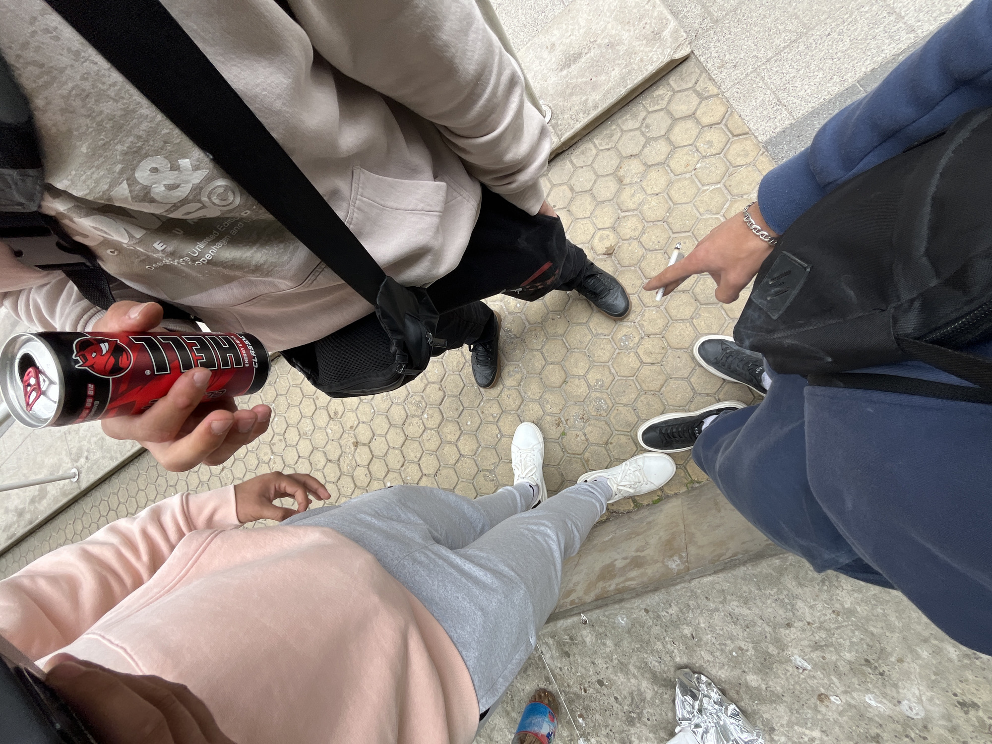Three individuals stand on a hexagonal-tiled surface in what appears to be an urban environment. From an overhead perspective, the person on the left wears a light pink hooded sweatshirt and gray sweatpants and holds a can of Hell energy drink. To their right, a person in a light beige hooded sweatshirt and black pants points toward a cigarette on the ground. The individual on the right wears a dark blue hooded sweatshirt and dark-colored shoes. The location is Plovdiv, Bulgaria.