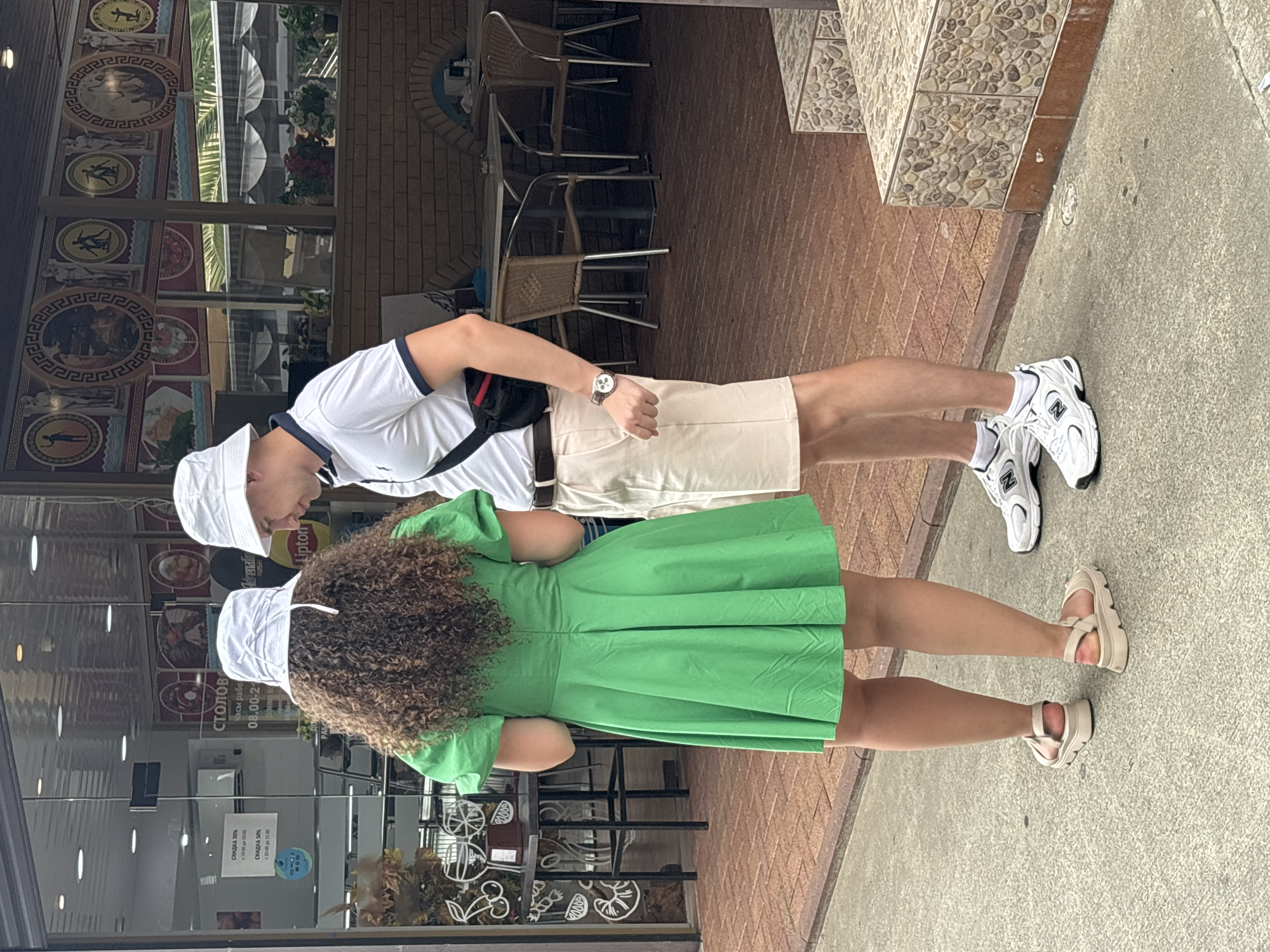 A man and a woman stand on a sidewalk in front of a restaurant or cafe with outdoor seating. The woman, on the left, has curly brown hair and wears a green dress and beige sandals. The man, on the right, wears a white polo shirt, beige shorts, white sneakers, and a white bucket hat. He has a black fanny pack worn across his chest. The background shows tables and chairs, as well as a building with decorative murals. The scene appears to be in Sochi, Russia, indicated by the text in Russian on a sign in the window, which mentions "столовая" (canteen/cafeteria) and working hours.