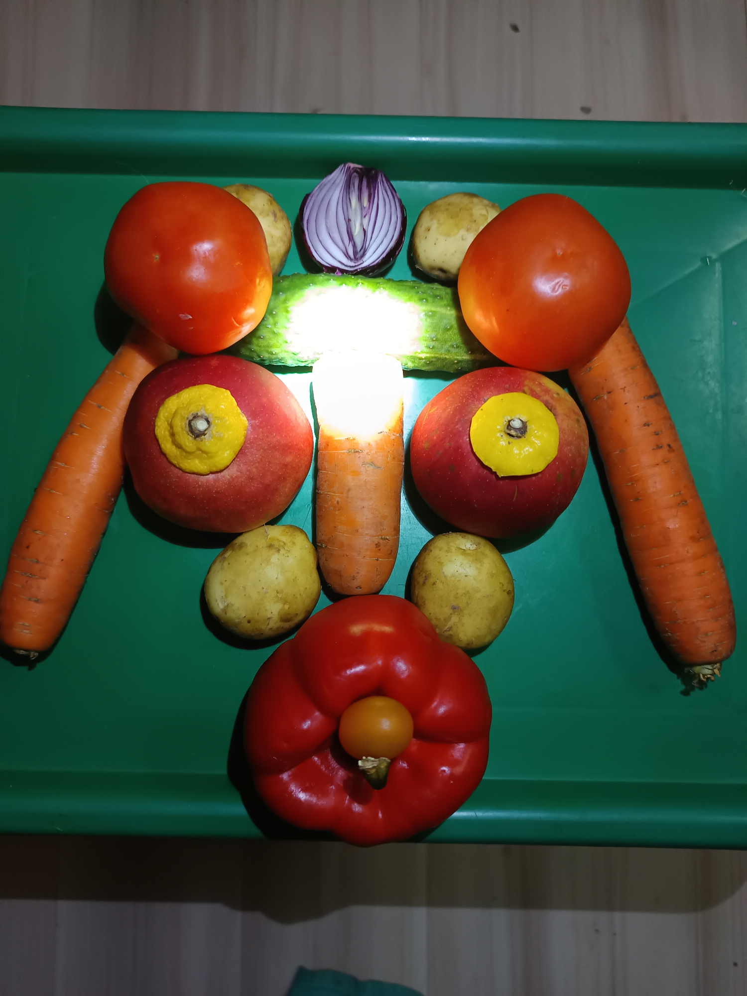 An overhead shot displays an arrangement of vegetables on a green plastic surface. The vegetables are arranged to resemble a face or a robot. From top to bottom, the arrangement includes a halved red onion, a cucumber, two tomatoes, two potatoes, two carrots positioned vertically on the sides, and a red bell pepper with a small tomato in its center. The lighting is bright, casting shadows. The background is out of focus and appears to be a wooden surface. The location in the city of Tyshky, Ukraine, is not discernible from the image.