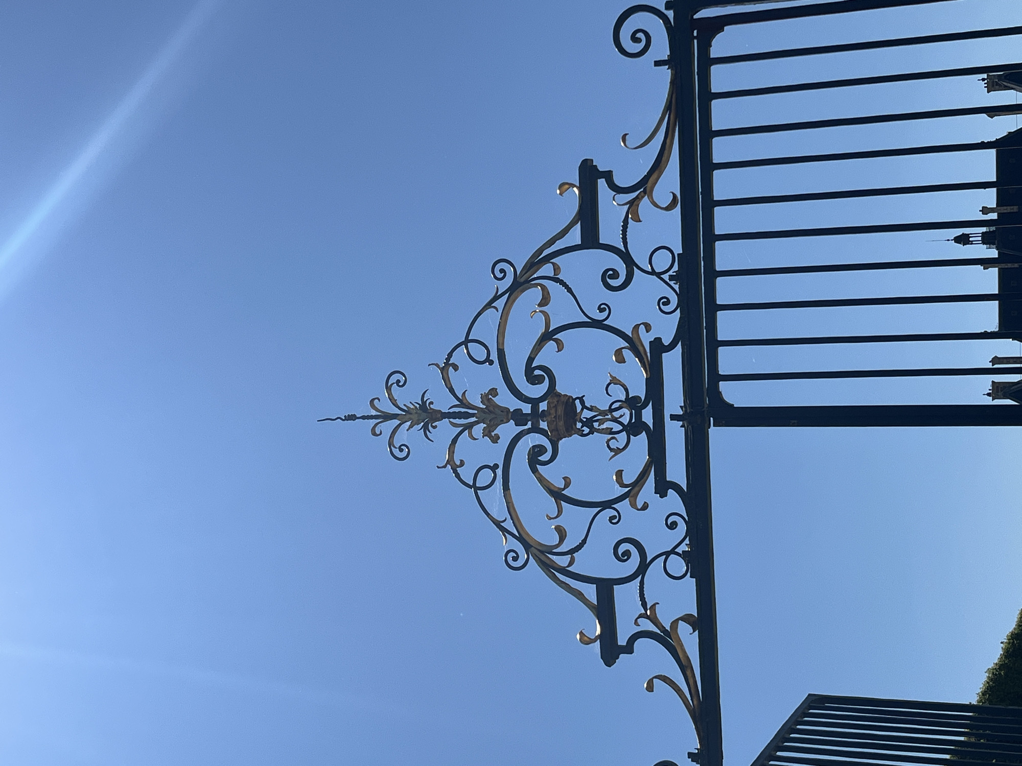 The image captures an ornate black and gold wrought iron gate against a clear blue sky. The gate features intricate scrollwork and a decorative crest at the top. Part of the gate is open, revealing a row of vertical bars. In the background, the roofline of a building with chimneys and a turret is visible. This scene is located in the city of Sceaux, France.