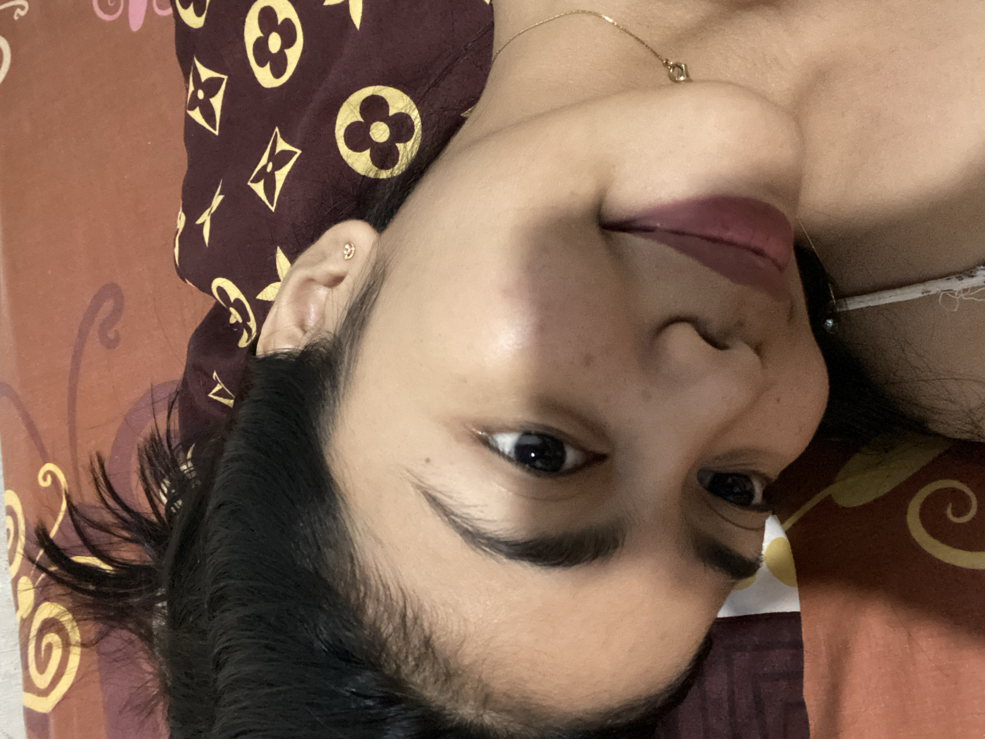 The image shows a close-up, tilted selfie of a young woman lying down, likely on a bed, as indicated by the patterned fabric. The woman has dark hair pulled back and is looking at the camera with a slight smile. She is wearing a small, delicate stud earring in her left ear and a thin gold necklace with a small pendant. The background fabric is a deep maroon color with a repeating pattern of yellow shapes, resembling stylized flowers and quatrefoils, possibly inspired by designer logos. The lighting suggests an indoor setting, possibly a bedroom in Madiun, Indonesia, given the context of the request, though no specific identifying landmarks are visible. The overall impression is a casual, personal moment captured in a photograph.