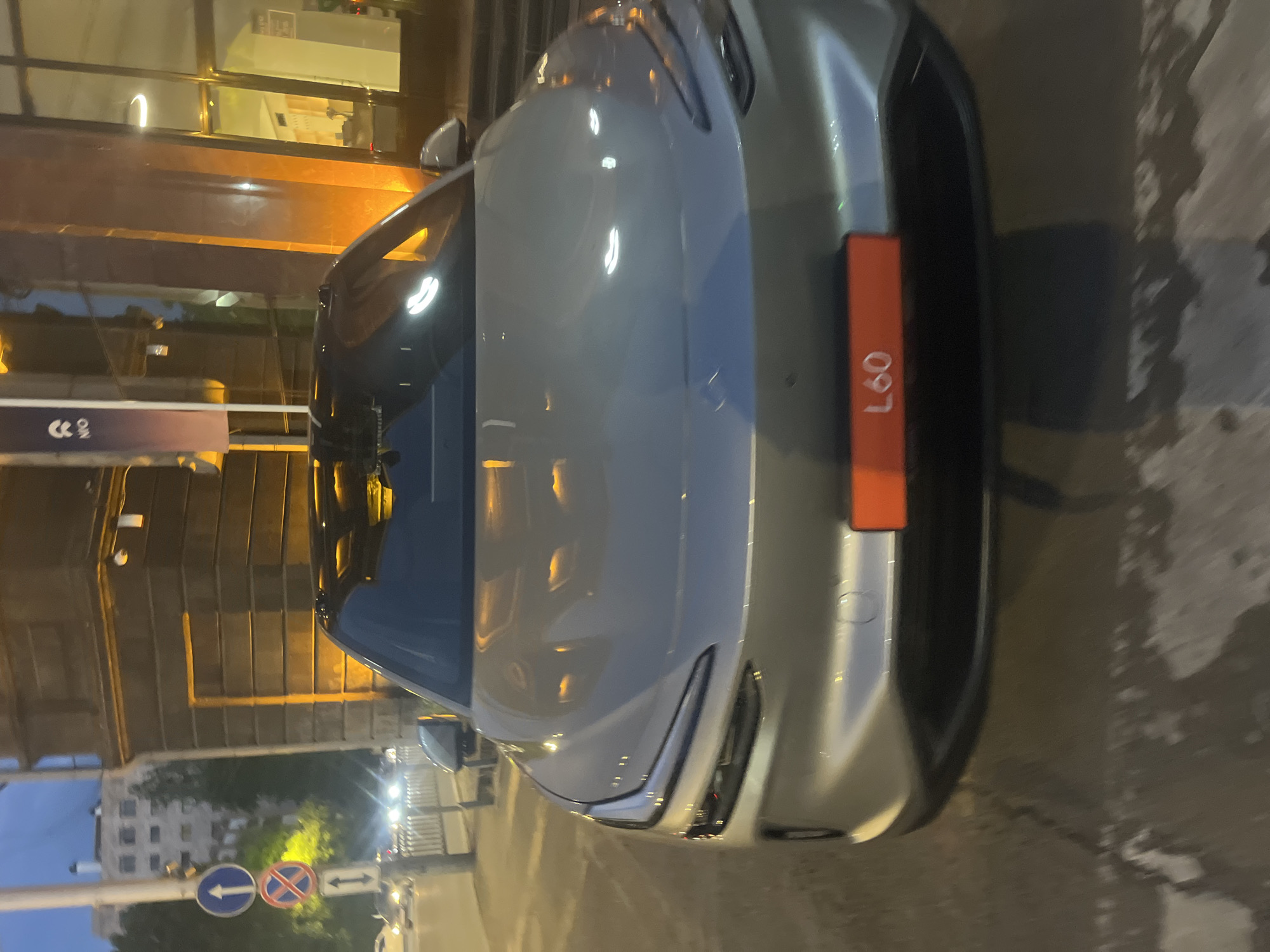 The image shows the front of a light-colored vehicle parked on a street in Yerevan, Armenia. The car has an orange sign on its front, displaying the alphanumeric code "L60". Traffic signs indicating lane directions are visible in the background. The reflection of the surrounding buildings and lights is apparent on the car's hood and windshield. The photograph was likely taken at dusk or night, given the low light conditions and artificial lighting.