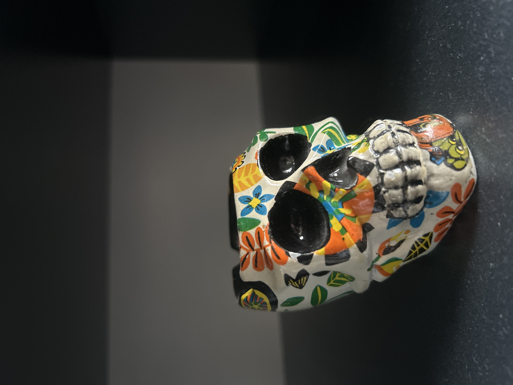 The image shows a colorful, decorative skull. It is white with floral and geometric patterns in shades of orange, blue, green, and yellow. The eye sockets are black. The skull appears to be ceramic or a similar material. The background is a dark gray with a gradient effect, suggesting a shelf or shadowed wall. The skull is located in Tbilisi, Georgia.