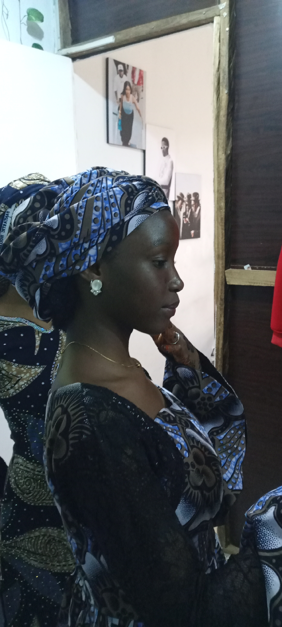 A woman in a blue patterned headscarf and dress stands indoors. There are framed photos on the wall behind her. The location is Unknown, Unknown.