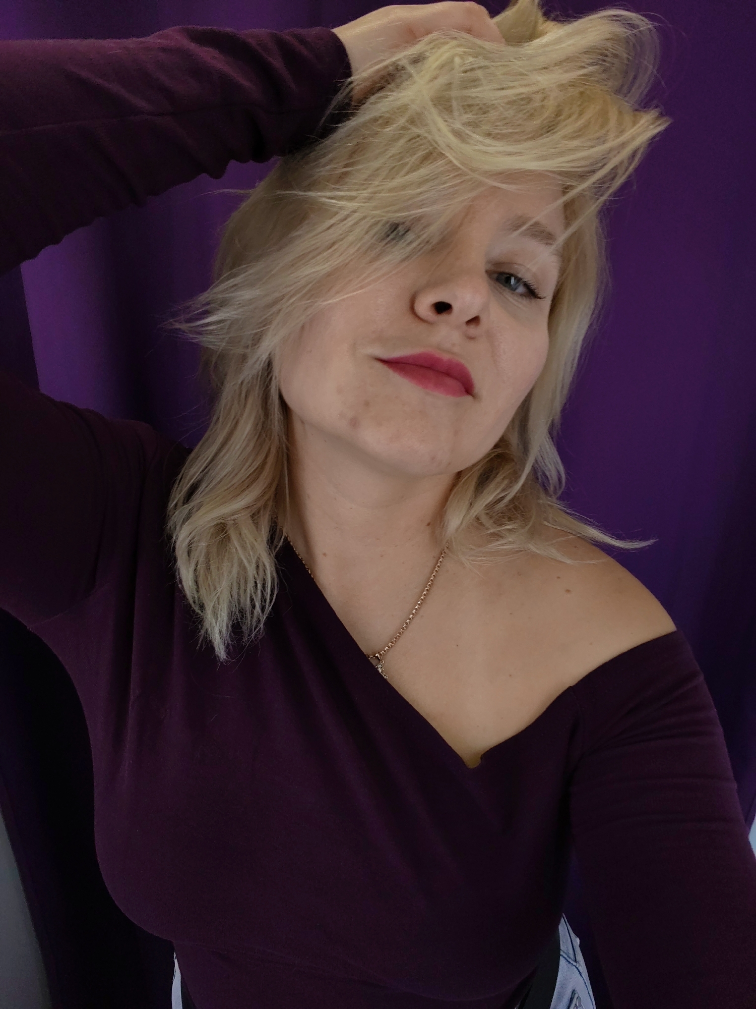 A woman with blonde hair and pink lipstick is posing for a selfie. She is wearing a dark purple, off-the-shoulder top and a gold necklace. Her left arm is raised, and she is running her fingers through her hair. The background is a purple curtain. The image appears to have been taken indoors, possibly in a home or hotel room. There is no information in the image to suggest it is located in Izhevsk, Russia.