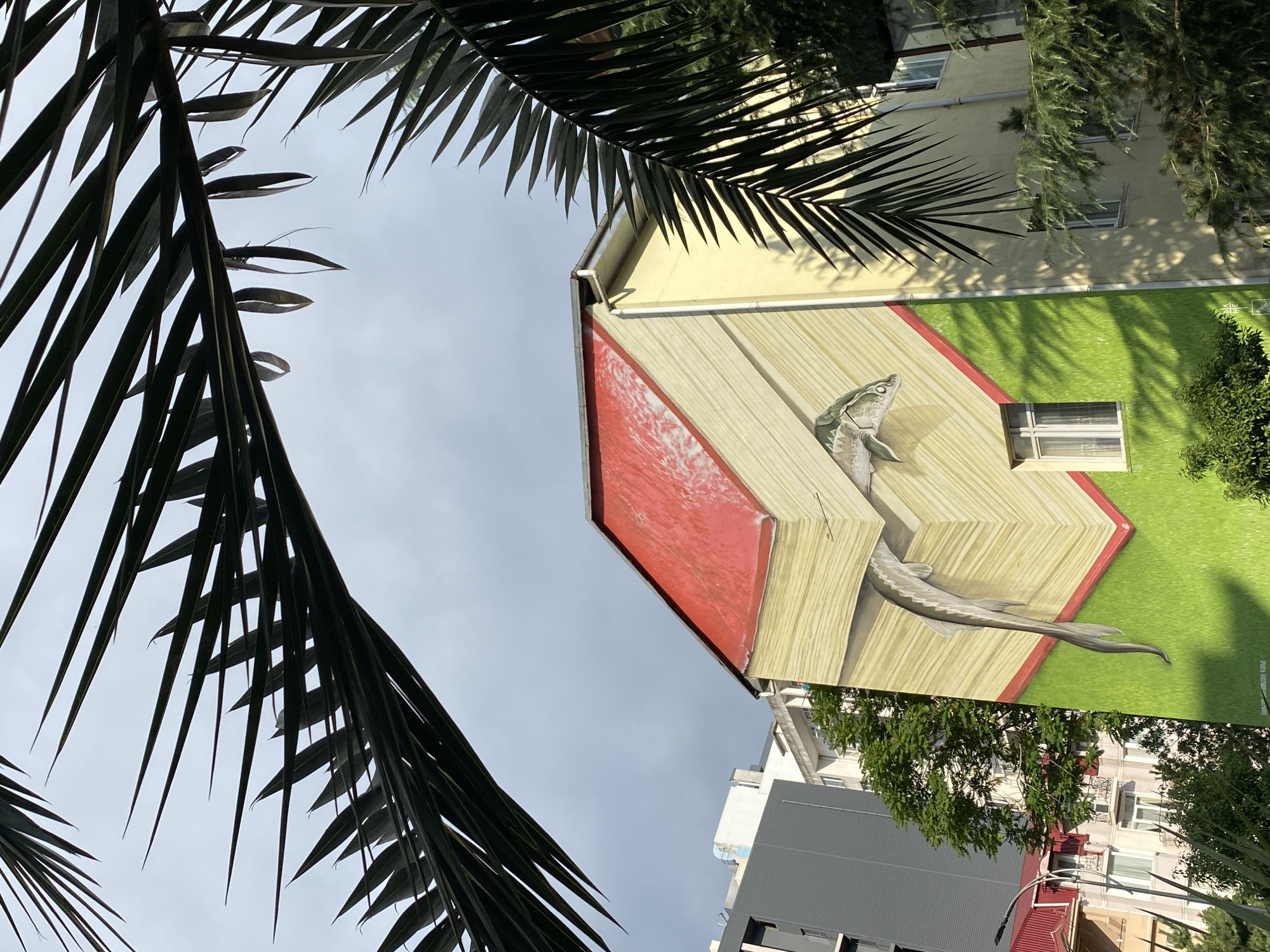 The image shows a building with a mural painted on its side, located in Batumi, Georgia. The mural depicts an open book with pages rendered in shades of yellow and brown, with a red cover. A sturgeon is painted emerging from the pages. The lower portion of the mural shows a bright green grass texture. Palm fronds are visible in the foreground, framing the scene against a cloudy sky. Other buildings, including a grey high-rise and a multi-story building with balconies, are visible behind the muraled building. Trees and foliage are also present. The mural is located on a light yellow building.