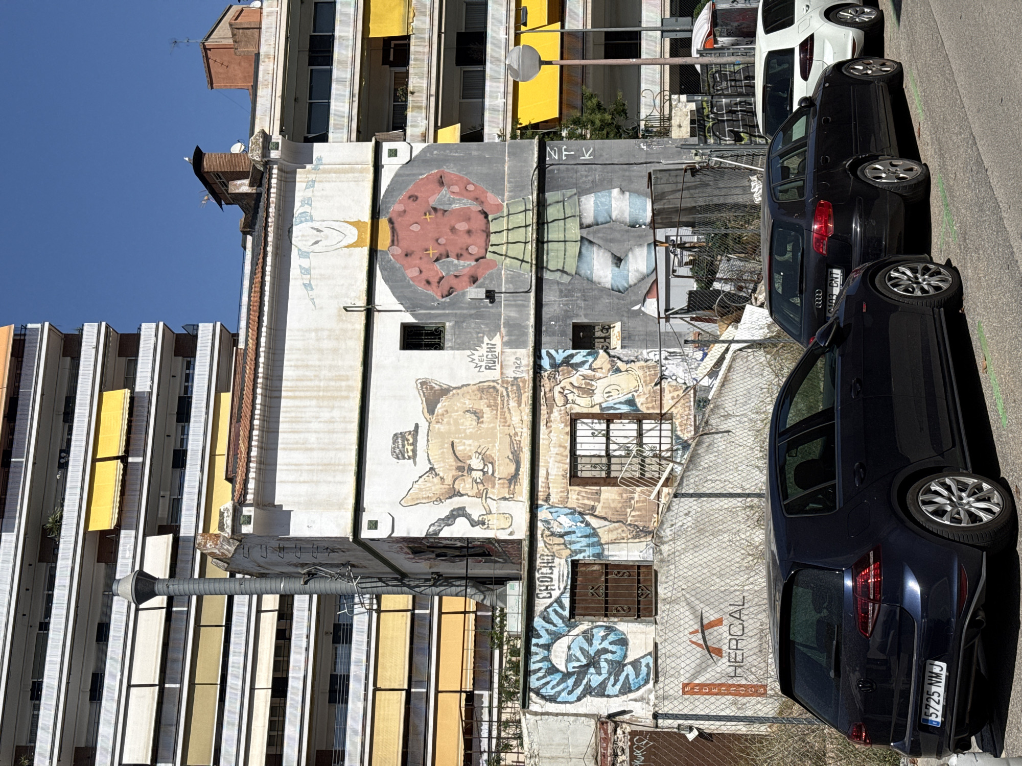The image shows a street scene in Barcelona, Spain, with a focus on a graffiti-covered building. In the foreground, three cars are parked on the street: a dark blue Volkswagen Golf on the left, a black Audi A3 in the middle, and a white Volkswagen Golf on the right. The building behind the cars features large murals. On the upper part of the building, a stylized cat wearing a hat and smoking a pipe is depicted. Below this, a large figure resembling a person with a yellow head and a red, polka-dotted torso is painted, wearing a green checkered skirt and striped pants. To the left of this figure, a blue, coiled serpent-like mural is visible. Several windows with metal bars are present on the building. In the background, a tall, multi-story apartment building with numerous balconies and yellow awnings stands out. A street lamp is visible on the right side of the image. The sky is clear and blue.