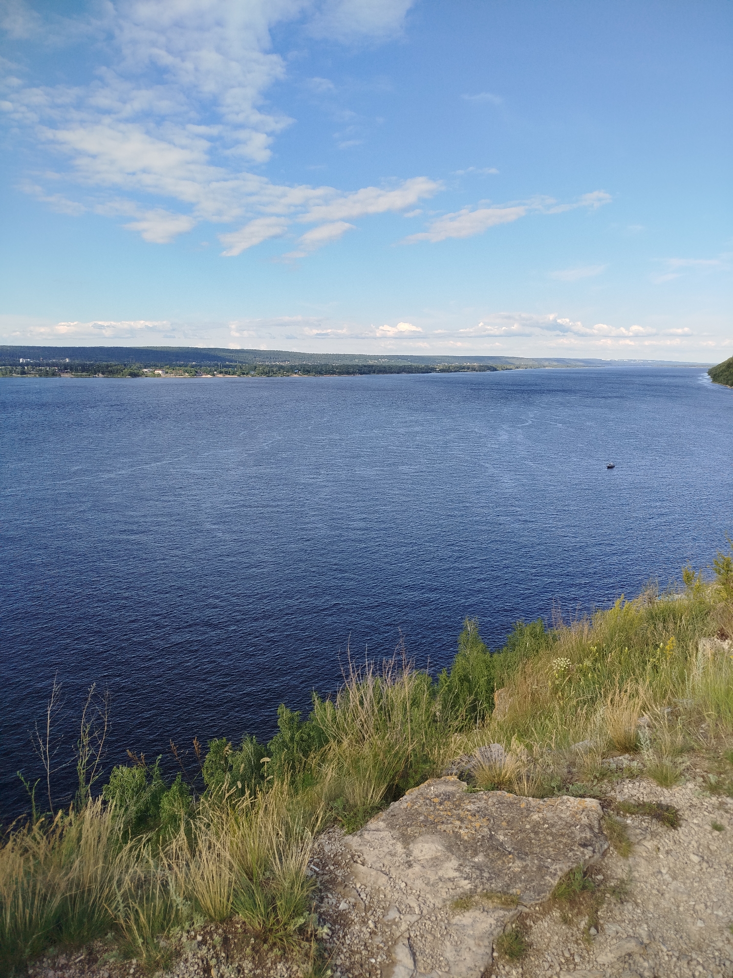 This media file captures a scenic vista of a wide, deep blue river under a bright, partly cloudy sky. The perspective is from a grassy, rocky cliff overlooking the water. In the foreground, dry grasses and rocky terrain dominate the cliff edge, suggesting a natural, elevated viewpoint. The river stretches out to the horizon, where a distant, hazy shoreline with trees and some buildings is visible. A small dark boat is present on the water, indicating a tranquil and possibly recreational setting.
The weather appears to be fair and sunny, with scattered white clouds in a clear blue sky. The lighting suggests it is daytime, likely late morning or early afternoon. The lush green vegetation on the cliff, contrasted with the dry grasses, hints at a temperate climate. The presence of a wide river and distant treed landscape is consistent with a natural park or rural area. Given the location context of Zhigulyovsk, Russia, this scene likely depicts the Volga River, a prominent geographical feature of the region known for its expansive width and picturesque surroundings. There is no visible text in the image. The overall mood conveyed is one of peace, natural beauty, and expansive vistas.