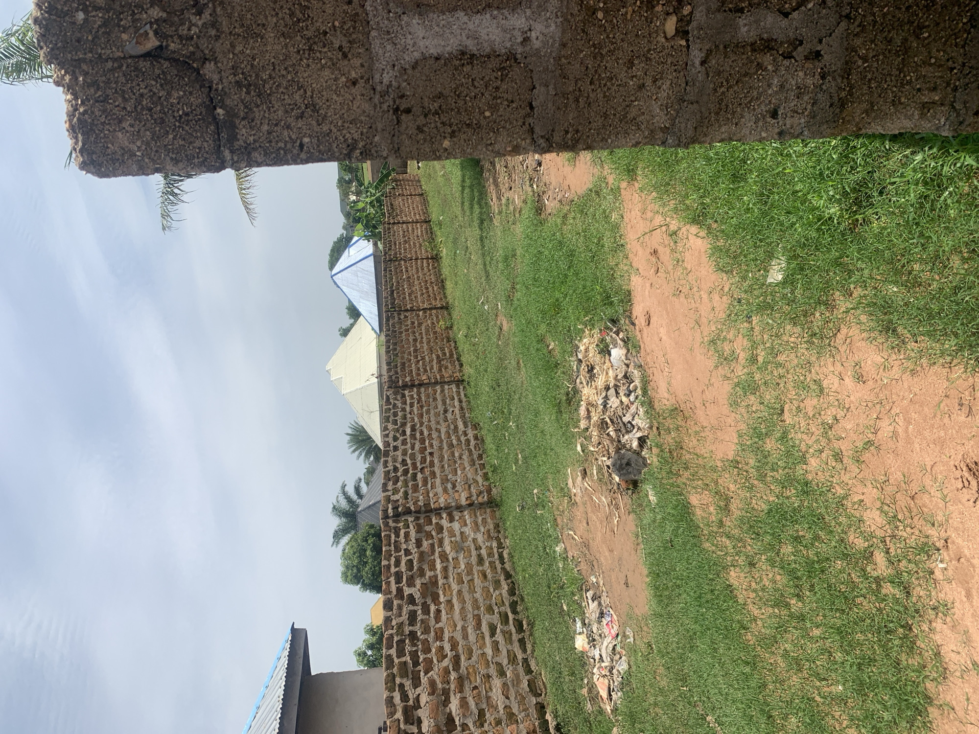 The image depicts a dusty plot of land bordered by a low, weathered brick wall. The wall is partially obscured by a rough, concrete pillar in the foreground, which appears to be part of a larger structure. Beyond the brick wall, a strip of sparse green grass lines the path, with patches of bare, reddish-brown earth interspersed. A pile of discarded trash, including plastic bags and debris, is visible near the wall on the left side of the image. In the background, several domed structures with metallic roofs are visible, along with scattered trees. The sky is overcast with light clouds. The scene suggests an undeveloped or partially developed area within a residential or semi-rural setting in Jauro Yinu, Nigeria, characterized by its unkempt appearance and evidence of construction and waste.