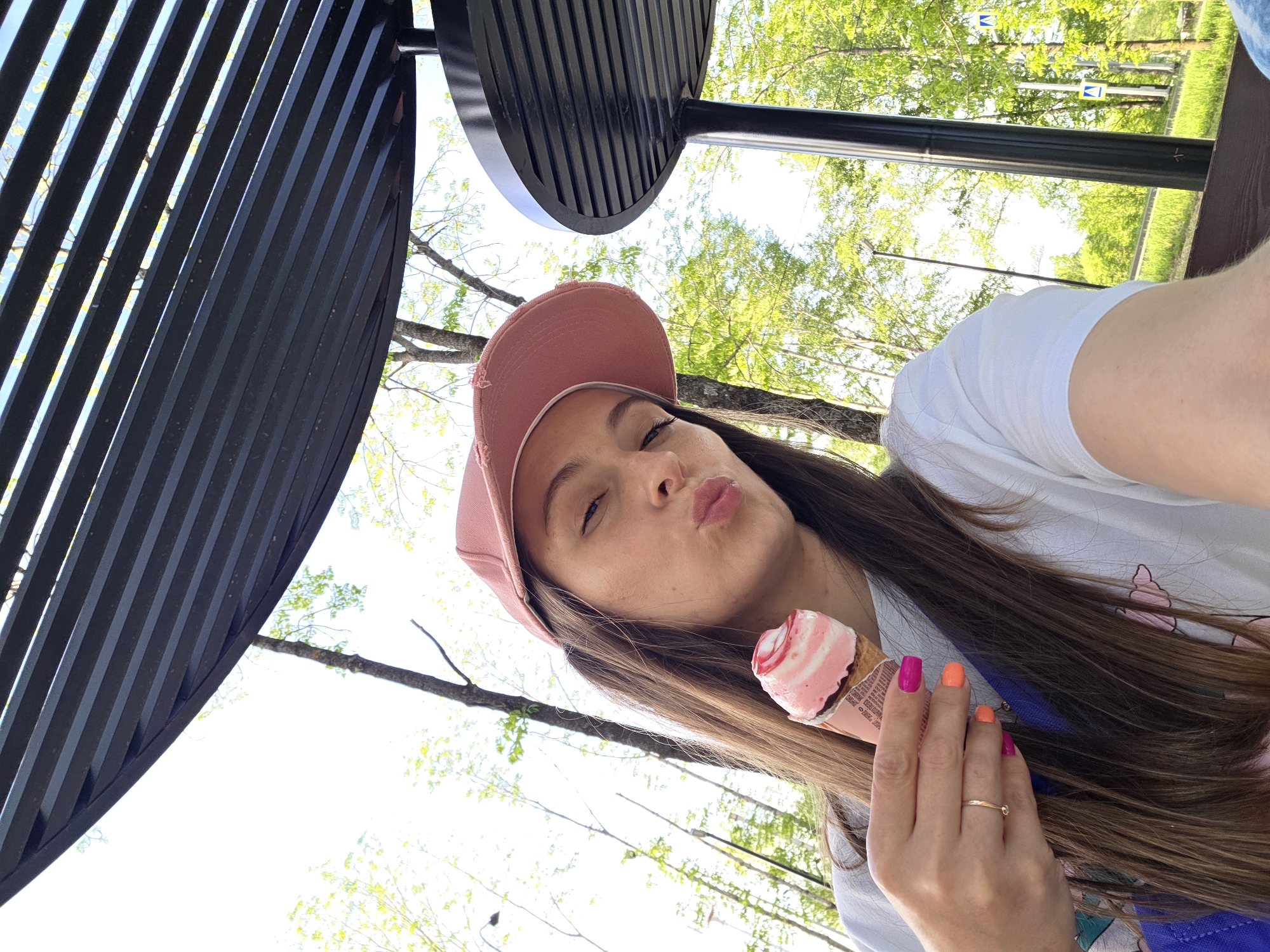 A woman holds an ice cream cone. She wears a pink hat and a white shirt. Her fingernails are painted with pink and orange polish. The background shows trees and a black metal structure. The location is Birobidzhan, Russia.