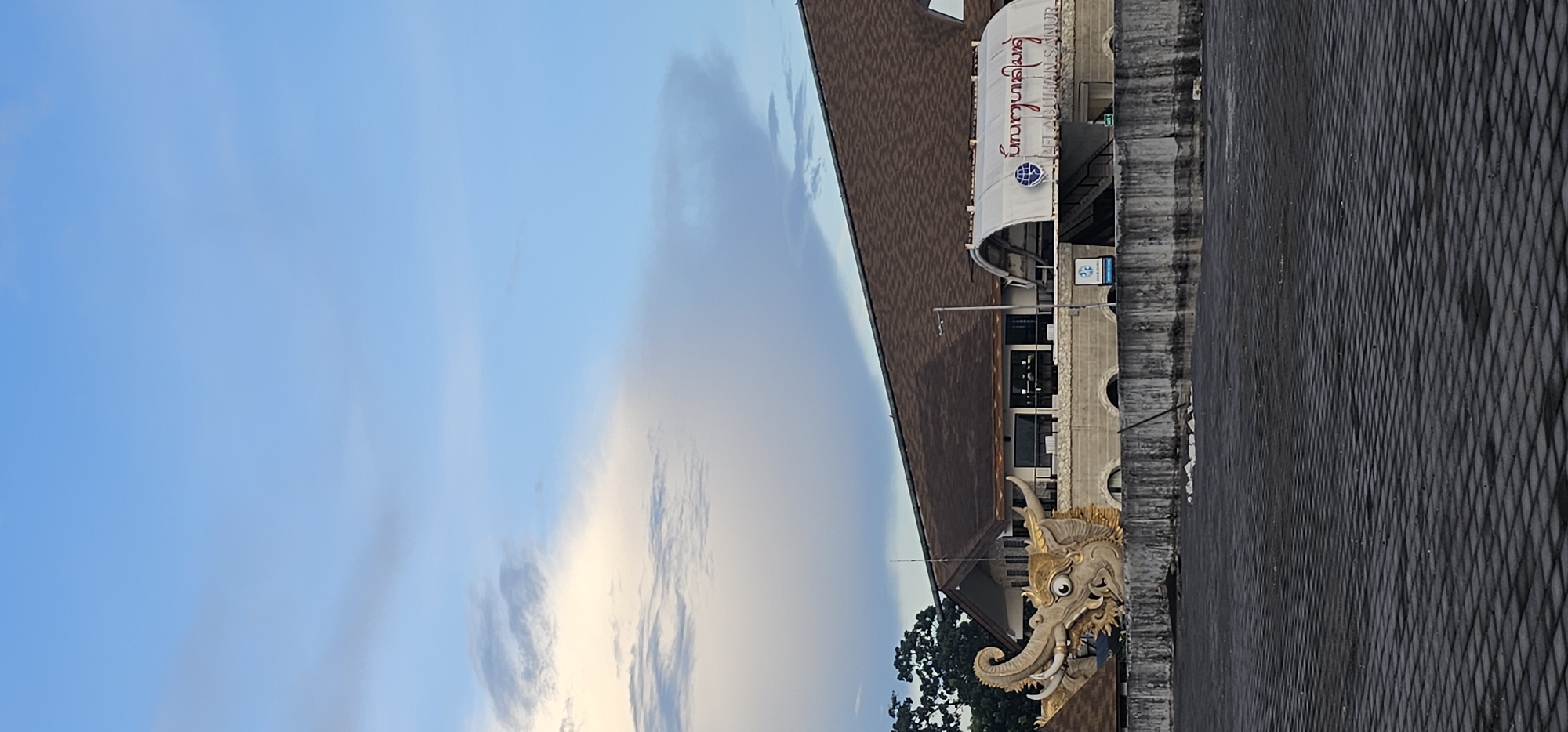 The image displays a static outdoor scene, likely captured during the soft light of early morning or late afternoon. A clear blue sky, sparsely populated with wispy, light-colored clouds, dominates the upper portion of the frame.
On the left, a multi-story building features a distinct dark brown, sloped roof. Extending from the building is a white awning, prominently displaying text. The text is in Thai script, reading "ธนาคารไทยพาณิชย์," which translates to "Siam Commercial Bank," and is also written in Romanized script as "SIAM COMMERCIAL BANK." A blue circular logo featuring a globe motif is positioned next to the text on the awning. Below the awning, the building shows dark windows and a textured facade that includes arched decorative openings.
A large, ornate sculpture of a golden elephant's head is integrated into the lower part of the building on the left. This highly detailed sculpture features tusks, large ears, and expressive eyes. Dark green foliage from trees can be seen behind the elephant sculpture and the lower building structure.
The right side of the image is occupied by a tall, dark gray wall or barrier with a pronounced diamond-patterned texture. This wall suggests a boundary or an adjacent structure. No human subjects or active events are depicted. While the text indicates "Siam Commercial Bank" (a Thai institution), the provided location context is Denpasar, Indonesia.