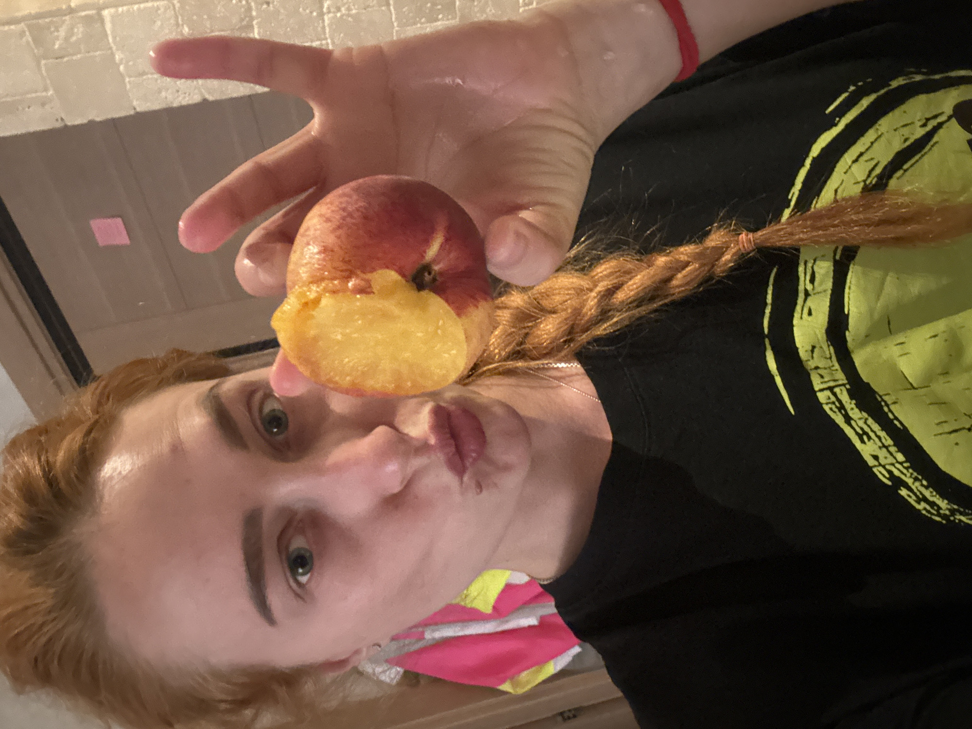 A young, fair-skinned person with reddish hair and green eyes holds a peach with a bite taken out of it in front of their face. They are wearing a black t-shirt with a graphic design in neon green. The person has a braid, and a red wristband is visible on their right wrist. The background shows a white tiled wall and a cabinet or drawer with a pink and white object on top. The location is not identifiable as Budva, Montenegro from the image.