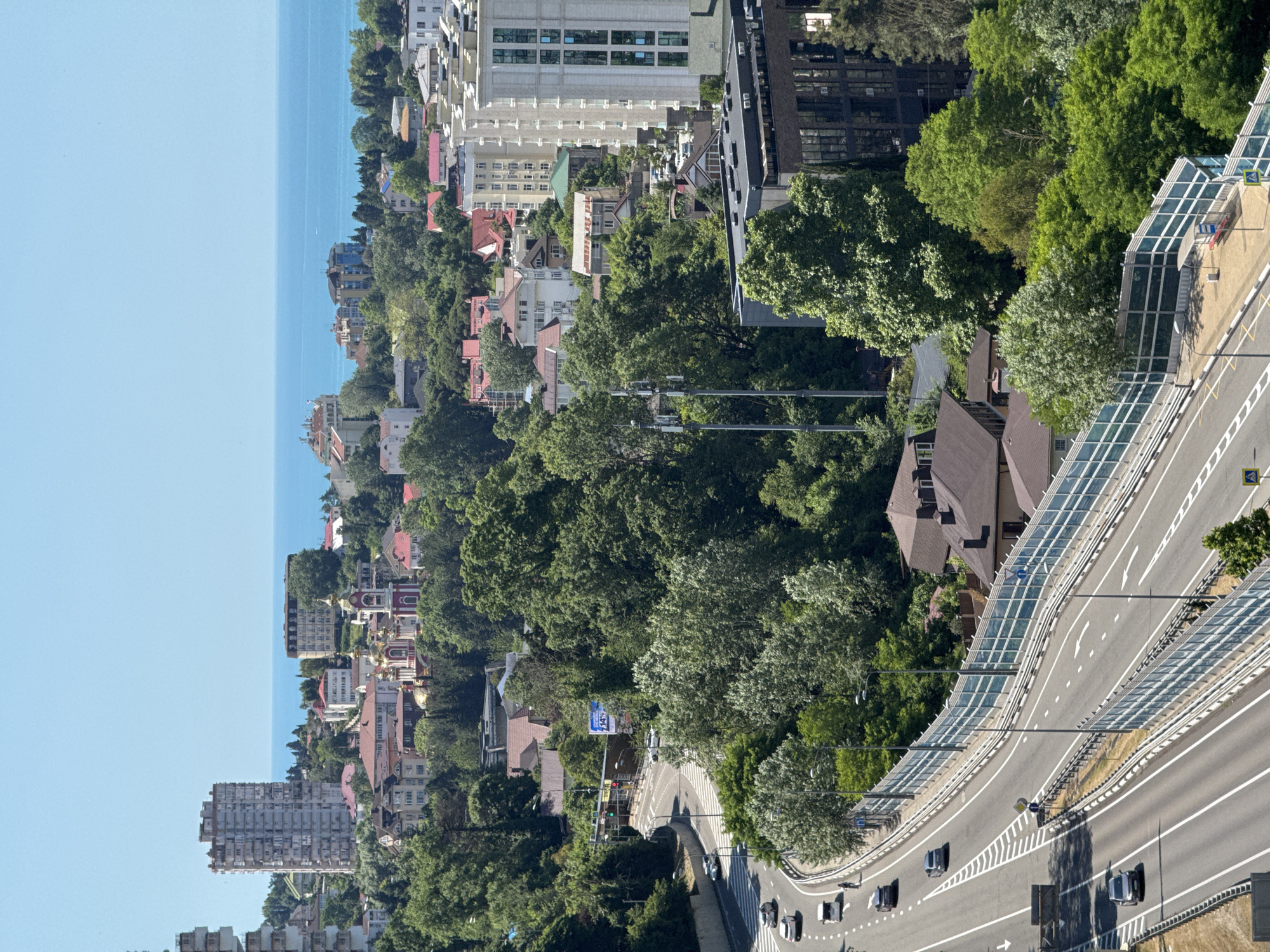 The image displays a cityscape with a focus on a multi-lane highway curving through a densely wooded area. Tall buildings, including a prominent high-rise apartment complex and a modern office building, are visible in the background, interspersed with red-tiled roofs of houses. A large body of water, likely the Black Sea, can be seen on the horizon. Several vehicles are traveling on the highway, and pedestrian walkways are present alongside it. There are also cell towers and streetlights along the road. The overall impression is a coastal city with significant urban development integrated into a lush, green environment. The scene is consistent with the urban landscape of Sochi, Russia, known for its coastal setting and mountainous terrain.