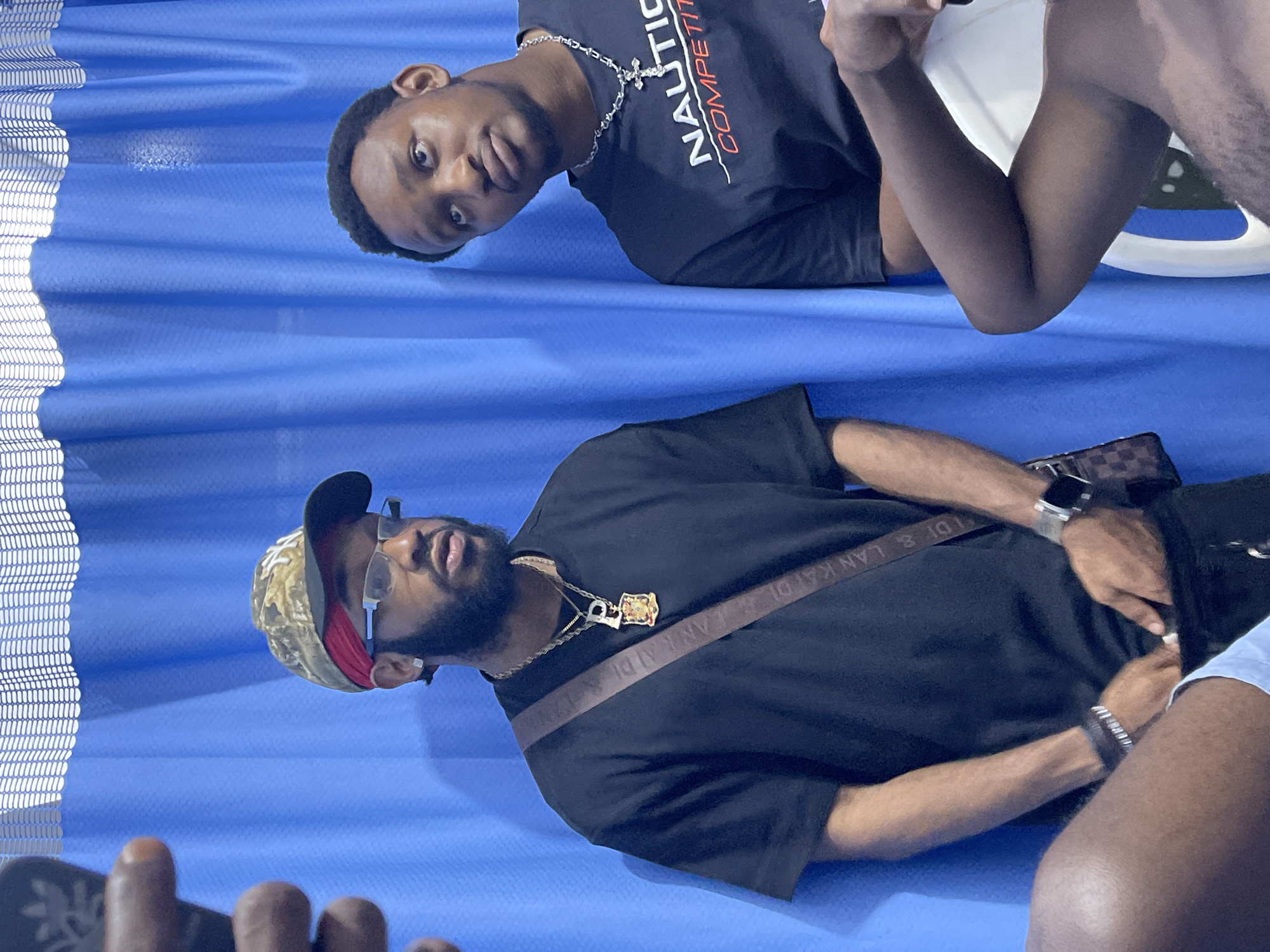 Two young men are standing in front of a bright blue curtain, likely in a public space in Abuja, Nigeria. The man on the left is wearing a camouflage baseball cap, a red bandana, glasses, a black t-shirt, and a crossbody bag. He has a beard and is wearing a chain with a pendant. The man on the right is wearing a black t-shirt with white and red text and a silver chain with a cross pendant. Both men appear to be looking in different directions, with the man on the right looking upwards. The blue curtain in the background has a textured pattern at the top. The lighting suggests an indoor environment, possibly a studio or a waiting area.