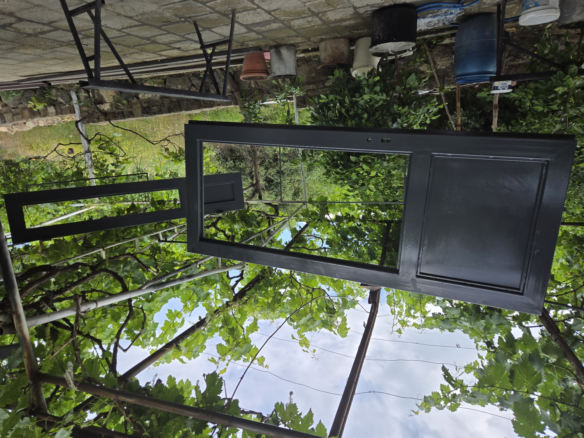 The image depicts an outdoor scene, likely a yard or garden, with an emphasis on a large, dark-colored, rectangular object that appears to be a door frame or a large panel. This object is positioned prominently in the foreground, dominating the central part of the frame. It is oriented such that its reflective surface shows a distorted view of the surrounding greenery and sky, suggesting it might be glass or a glossy finish.
Behind the door frame, lush green foliage from trees or vines is visible, creating a natural backdrop. Some of the branches and leaves are quite dense, partially obscuring what lies beyond. In the upper background, a stone wall can be seen, and further back, several buckets or containers are placed on a surface that looks like a tiled or paved area. These containers appear to be of various sizes and colors, including some dark ones, a terracotta pot, and a blue barrel. A metal structure, possibly part of a frame or support system, is also present, extending from the top left of the image.
The sky is visible through the gaps in the foliage and the open sections of the door frame, indicating it is daytime. The lighting suggests it could be a bright, possibly sunny day, though clouds are also present. The overall impression is of an outdoor work or storage area where a door or similar item is being handled or stored amidst natural elements. No people are visible in the image. No discernible text is present.
