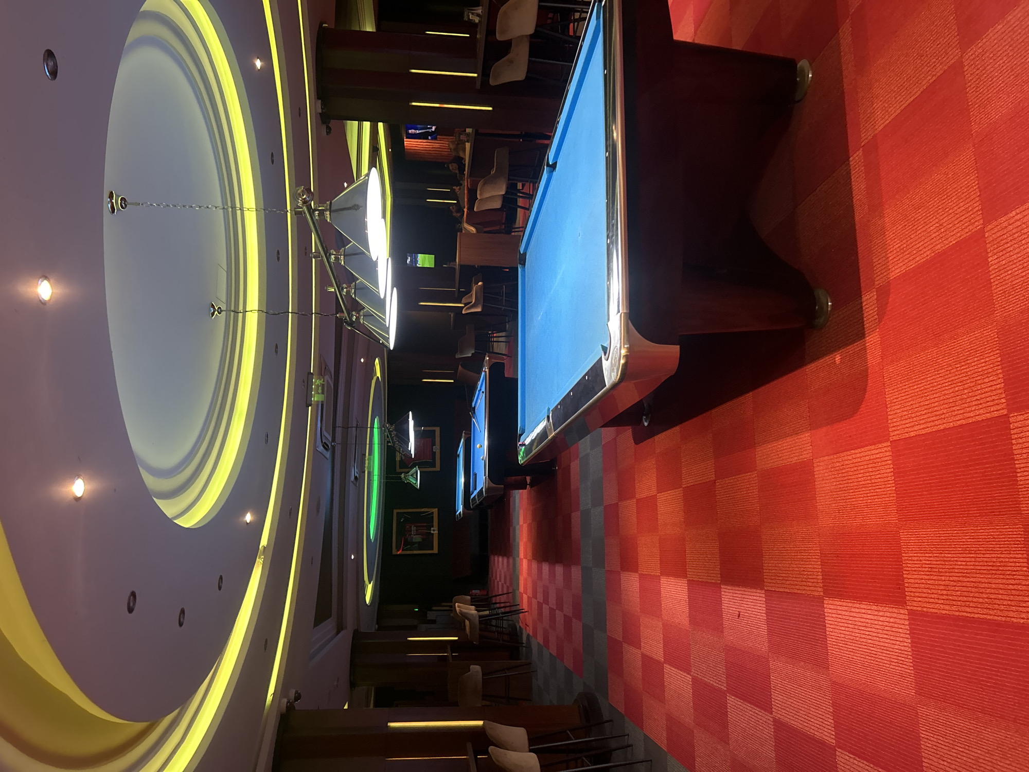 The image shows the interior of a billiard club in Sofia, Bulgaria. There are several pool tables with blue felt visible, each illuminated by a rectangular overhead light fixture. The flooring is a red and gray checkered pattern. Chairs and bar stools are arranged along a counter behind the pool tables. The ceiling features concentric circles of recessed lighting. Several pictures and screens are seen along the back wall. No people are clearly visible in the image.
