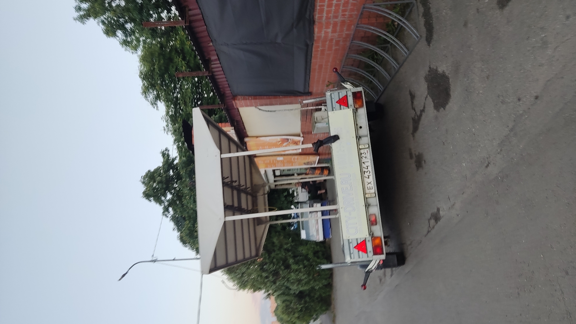 The image shows a trailer parked on an asphalt surface next to a brick building with a dark tarp covering part of its facade. Green trees are visible in the background, and the sky is overcast, suggesting it might be a cloudy day. The trailer has a white roof and a metal frame. There is some visible text on the back of the trailer, including what appears to be a license plate with the characters "EX 4341 23" and "CITY-DRIVER.RU". A person can be seen through the trailer's opening, and there are various items inside and around the trailer. The scene appears to be outdoors, possibly in an urban or industrial area in Krasnodar, Russia, given the location context. The overall mood is neutral.