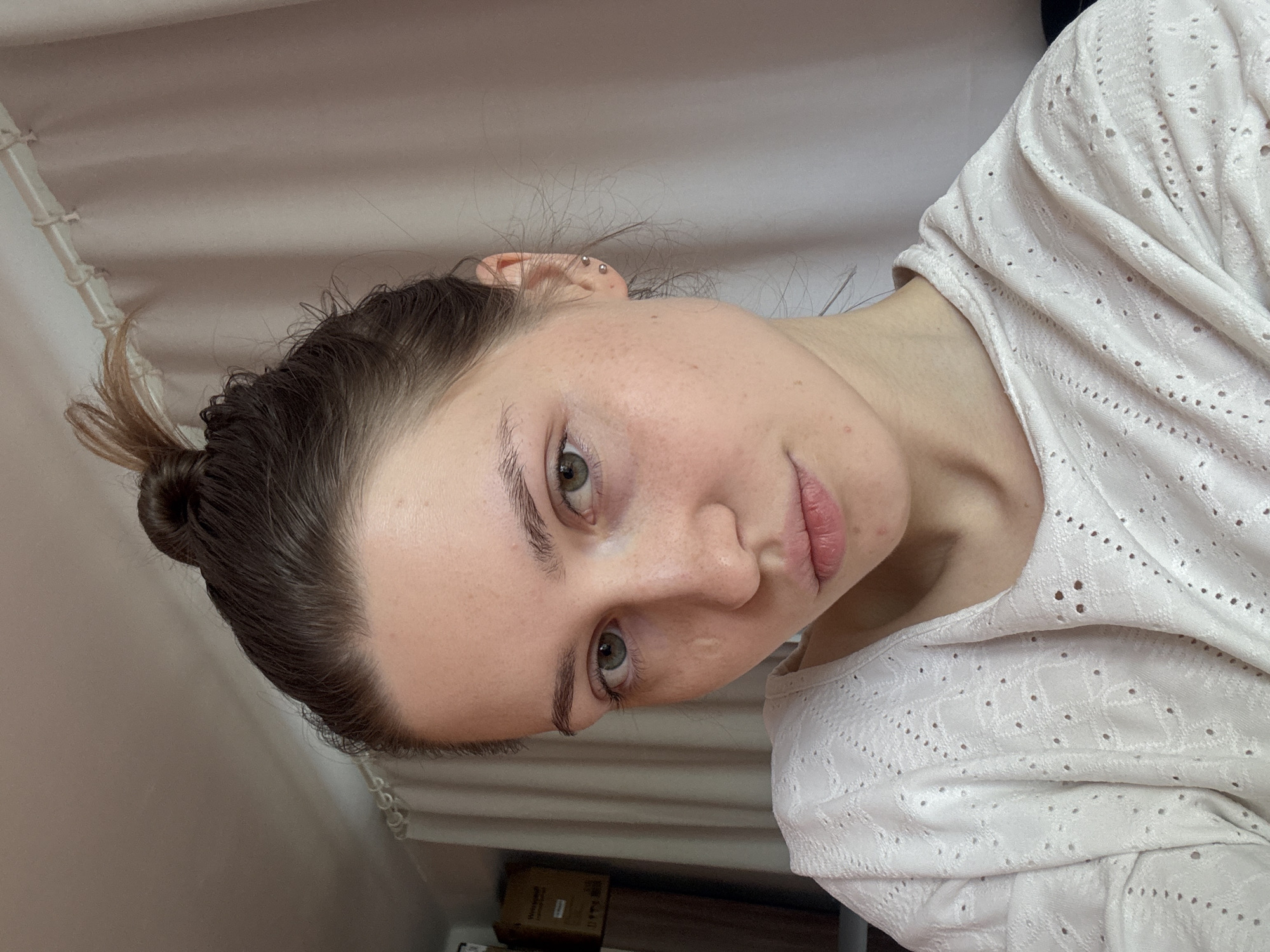 A woman with fair skin and light brown hair styled in a bun is facing the camera with a slight tilt. She has green eyes and is wearing a white lace-patterned top. The background is blurry but appears to be a neutral-colored wall. There is no indication of Saint Petersburg, Russia.
