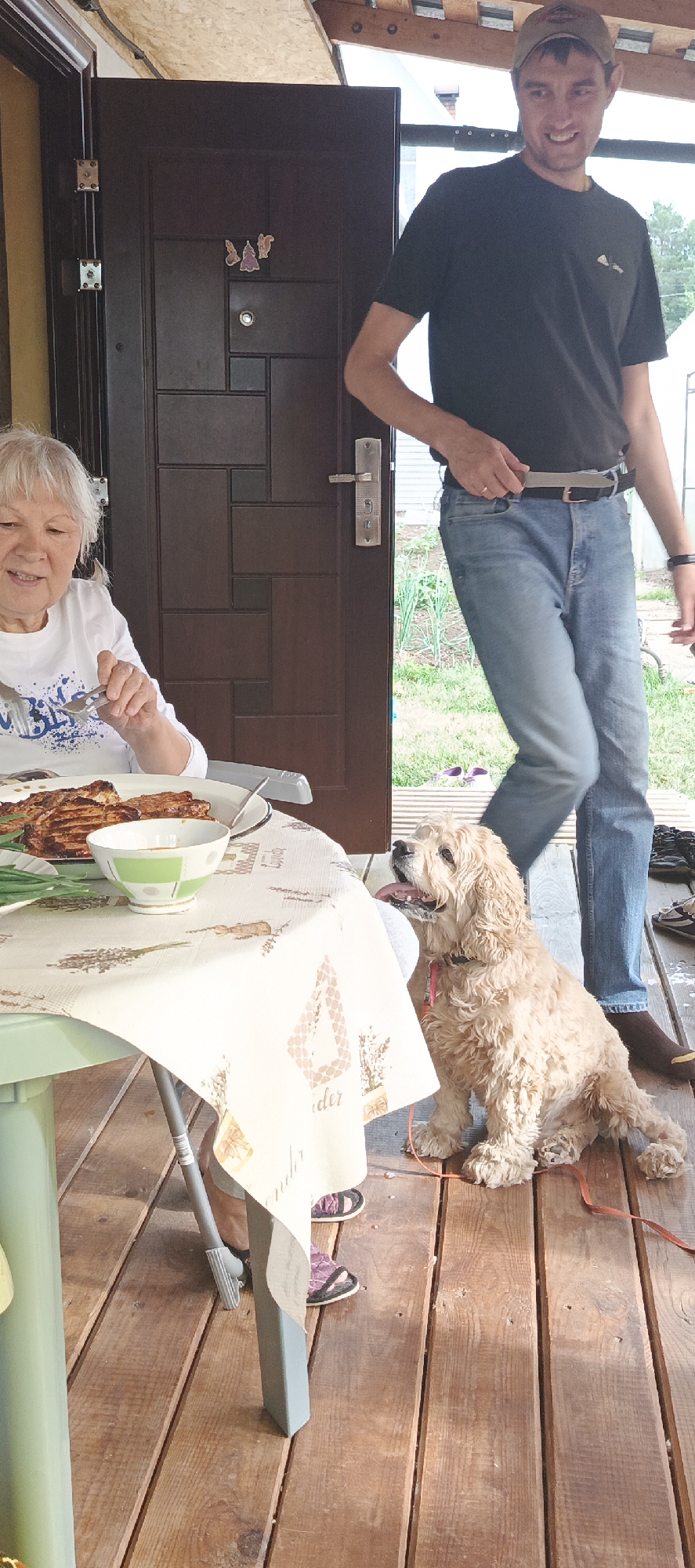 In Chaikovsky, Russia, a woman with white hair sits at a table on a wooden porch, eating a meal of grilled meat and what appears to be a side salad or vegetables. She is wearing a white t-shirt with blue lettering. To her right, a light brown dog with curly fur sits expectantly, looking up and to the side with its tongue slightly out. A man, wearing a dark t-shirt, blue jeans, and a baseball cap, stands to the right of the woman and behind the dog. He is smiling and looking off to the side. The wooden door behind them suggests they are at a house. The table is covered with a tablecloth patterned with lavender sprigs and geometric shapes. The background shows greenery, indicating an outdoor setting.