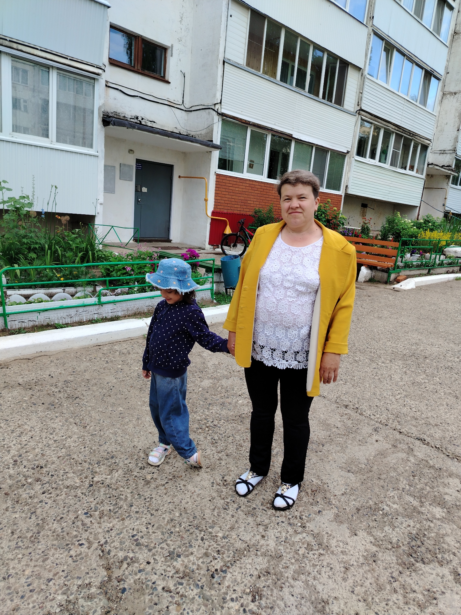 A woman and a young child stand hand-in-hand on a paved area in front of a multi-story apartment building. The building, likely in Noviy, Russia, has a white facade with numerous windows, some of which are balconies with white railings. A section of the building is bricked, and a yellow gas pipe runs vertically along the wall. Green metal fencing borders a flower bed with lush greenery and colorful flowers. A wooden bench is visible near the flower bed, and a bicycle and blue bin are to the left of the bench.
The woman, wearing a bright yellow jacket over a white lace top and black pants, looks towards the camera with a neutral expression. The child, dressed in a dark blue polka-dot top, jeans, and a blue bucket hat, looks down and to the side, her hand held by the woman. Both are wearing sandals. The ground is concrete with visible gravel. The lighting suggests it is daytime, with overcast conditions. The overall scene depicts a casual moment outside a residential building. No visible text is present in the image.