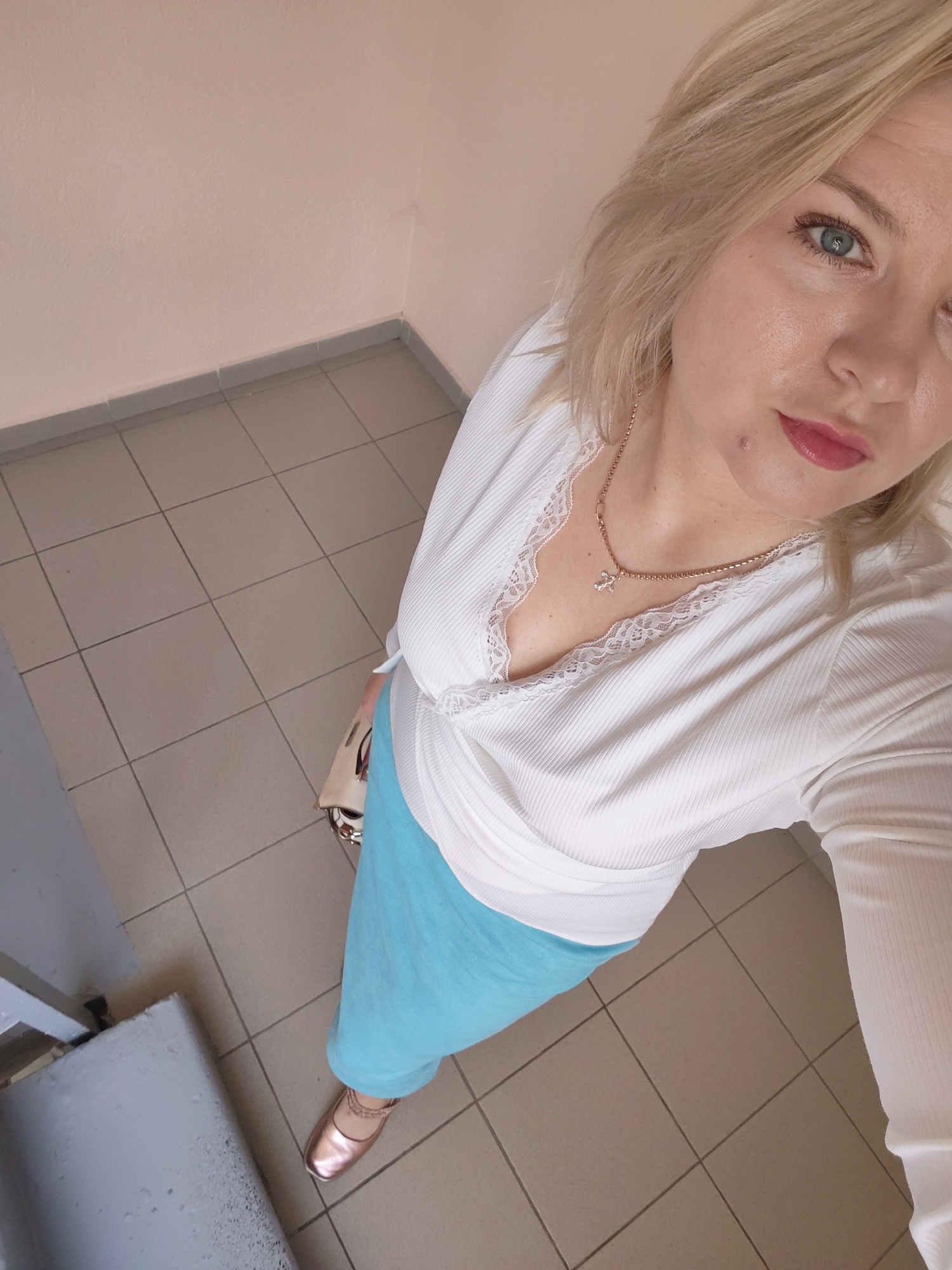 A high-angle shot captures a woman in a white ribbed top with lace trim and a light blue skirt. She is wearing rose gold ballet flats and a rose gold chain necklace with a cross pendant. A portion of a light-colored handbag is visible. The background consists of tiled flooring and a light-colored wall. The scene appears to be indoors, possibly in a hallway or waiting area. The lighting suggests daytime. The woman's expression is neutral. No visible text is present in the image.