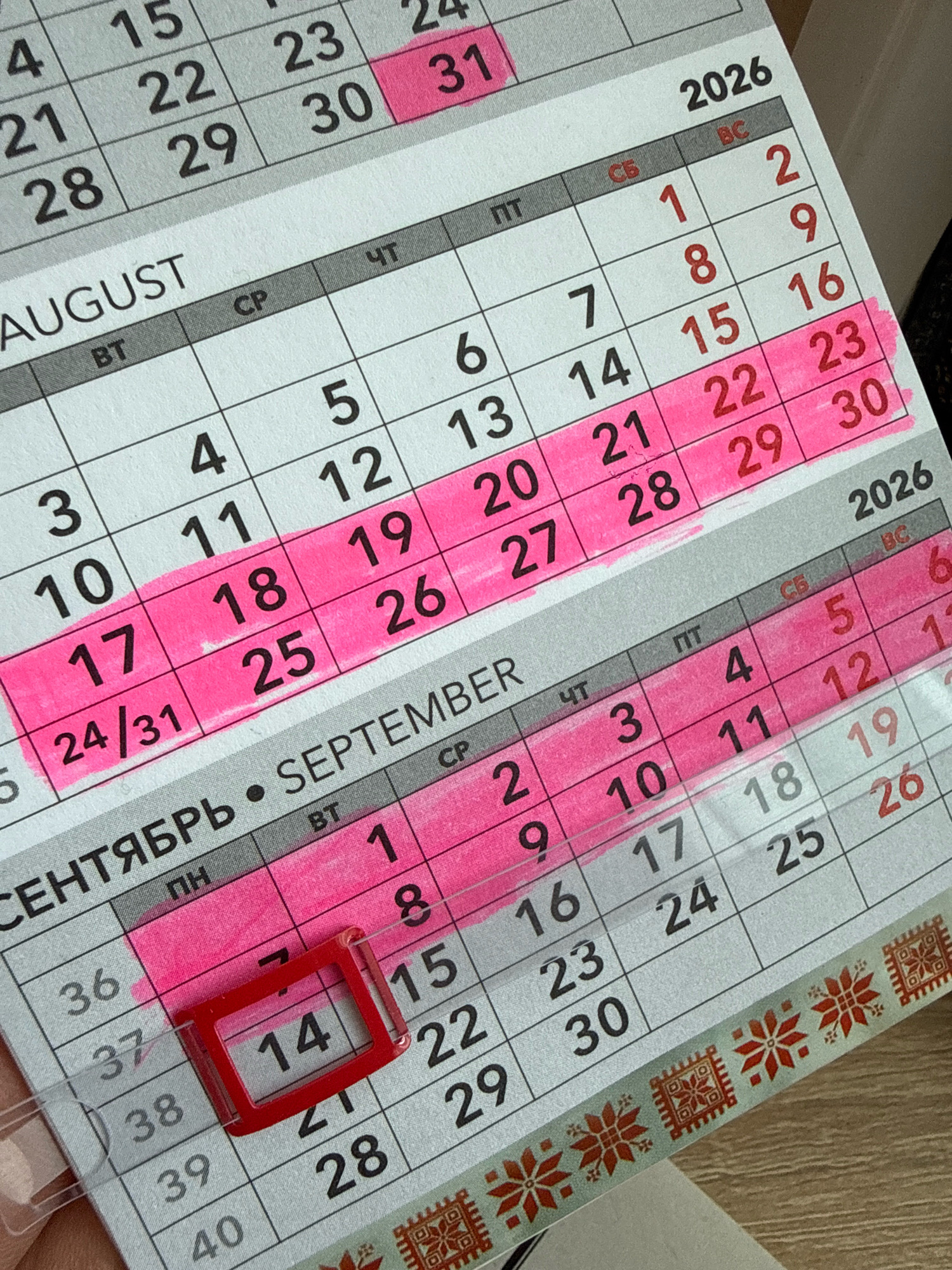 The image shows a close-up of a calendar for the year 2026, specifically featuring the months of August and September. The calendar is laid out in a grid format with days of the week labeled at the top. Some dates are highlighted with pink marker. A small red rectangular object, possibly a SIM card ejector tool, is placed over the date "14" in September. Decorative washi tape is visible along the bottom edge of the calendar. The background suggests a wooden surface. The calendar is positioned at a slight angle. There is no indication of the location being Slawharad, Belarus, or any other specific city.