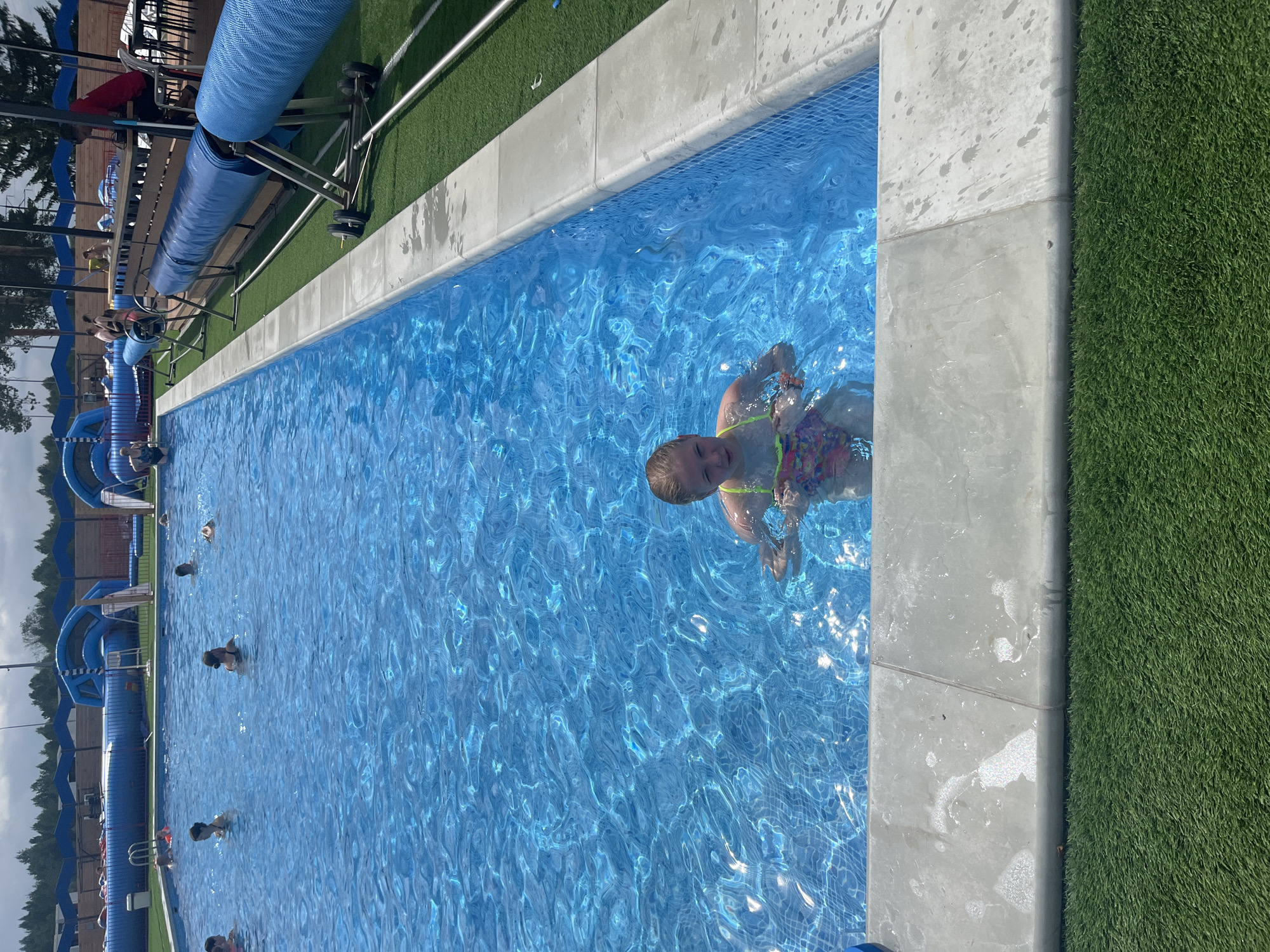 A young girl with blonde hair is swimming in a bright blue swimming pool. She is wearing a colorful swimsuit and is looking towards the camera with a slight smile. The water is clear and reflects the sunlight, creating a shimmering effect. The pool is surrounded by a concrete edge, and artificial turf lines the area next to the pool. In the background, there are inflatable structures that appear to be part of a water park, along with trees and a cloudy sky, suggesting it is daytime. Several other people, including children and adults, are visible in the pool, some of whom appear to be playing or relaxing in the water. The overall scene depicts a leisurely day at a swimming facility. The location context is Votkinsk, Russia.